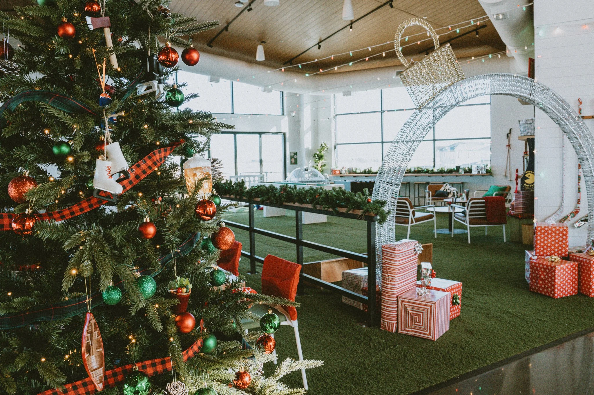 Decorated Christmas tree with red and green ornaments and plaid ribbon in a bright, modern room with large windows, string lights, and wrapped gifts underneath.