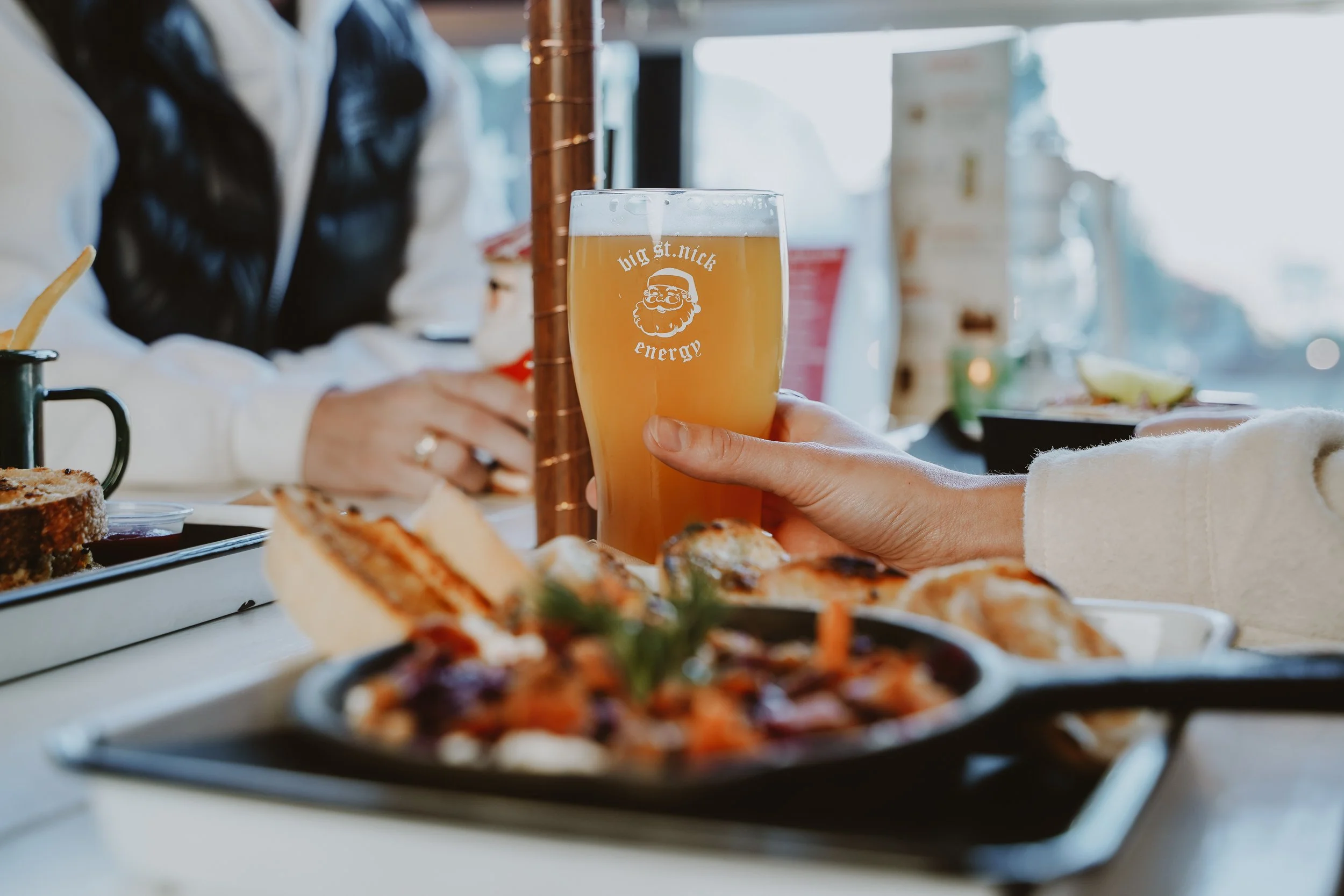 A person holding a glass of beer with a Santa hat logo and the text 'big st. nick energy.' In the background, there are plates of pizza, bread, and other food, with a blurred restaurant setting.