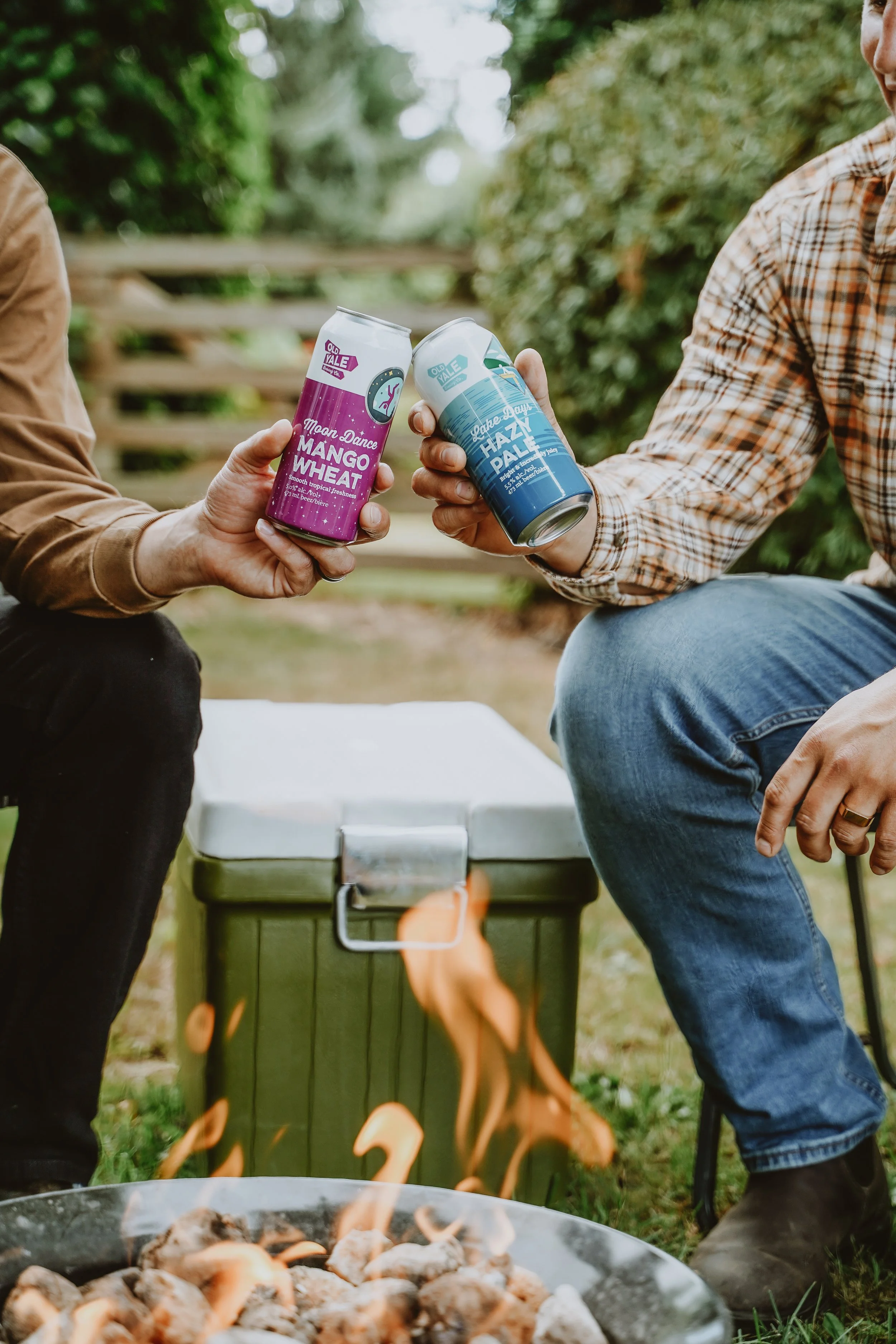 Two people toasting with cans of Old Yale beer near a small outdoor fire, with a green cooler and greenery in the background.
