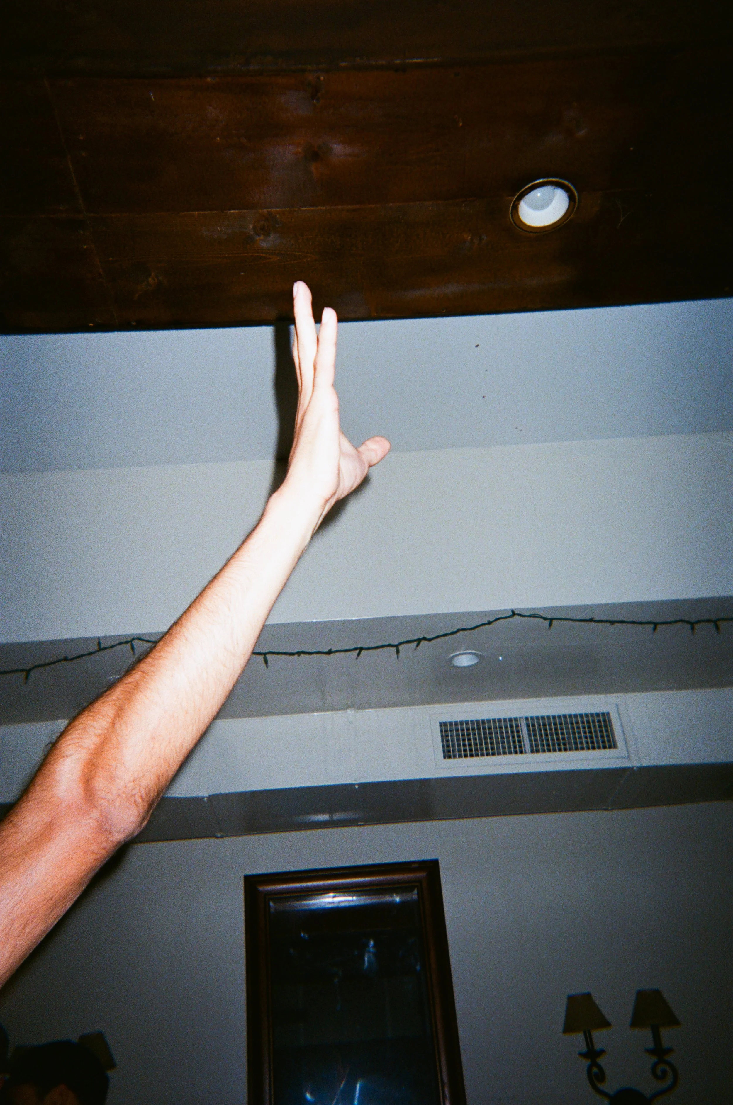 A hand reaching upward towards a wooden ceiling with a recessed light fixture, ceiling vents, and wall decorations.