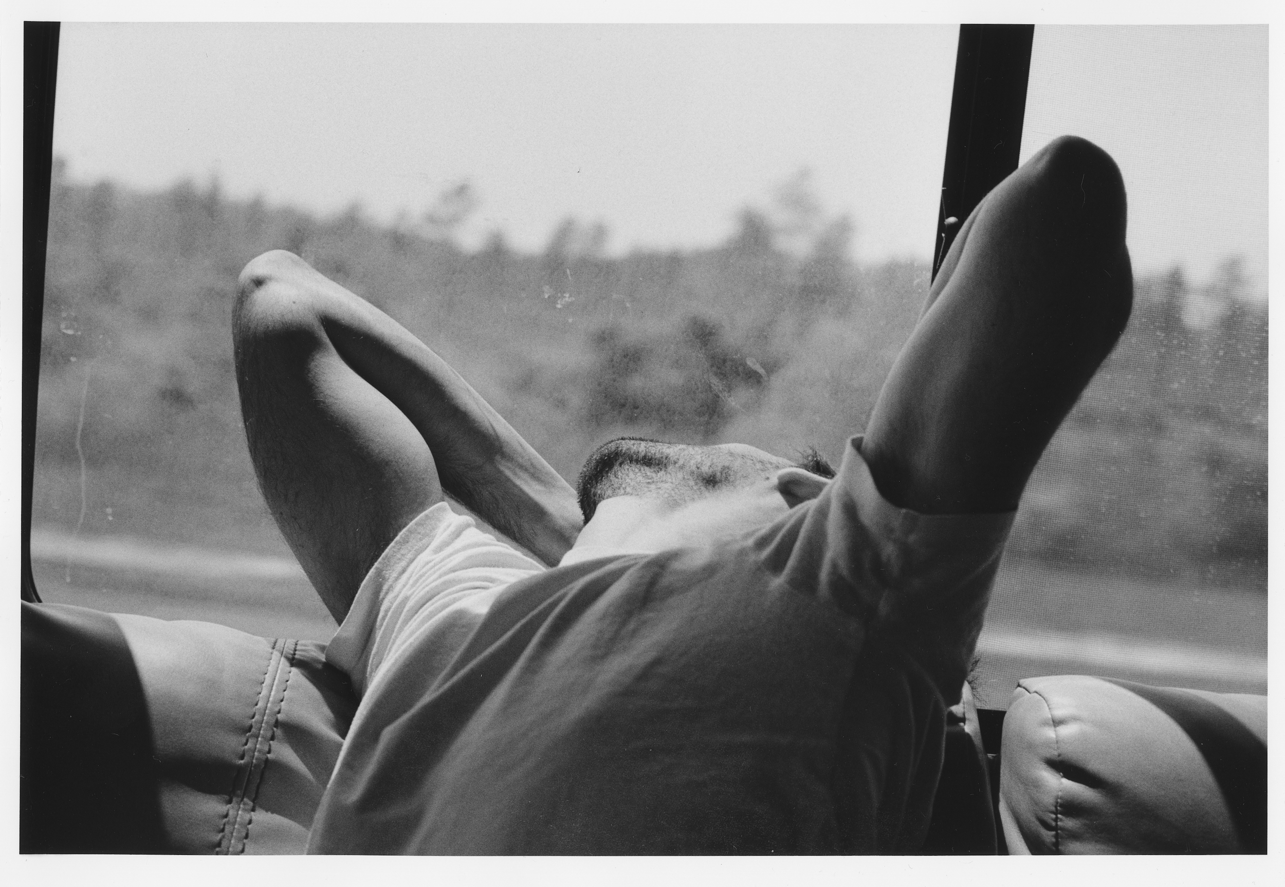 Black and white photograph of a person lying down, with legs raised and hands behind their head, in an airplane or car with mountains and sky visible through the window.