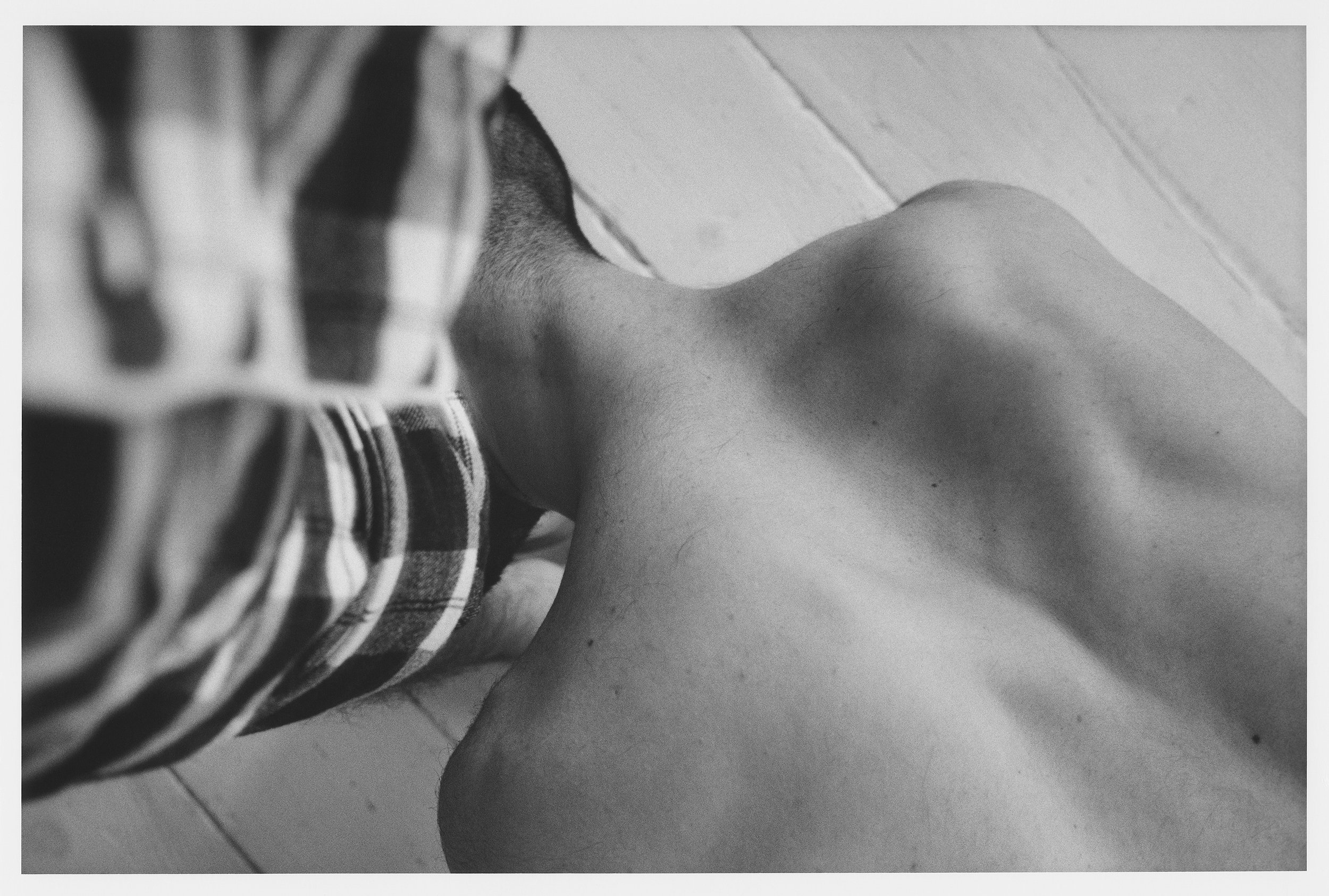 Black and white photo showing a person's leg and foot, wearing a plaid shirt, next to a tiled floor.