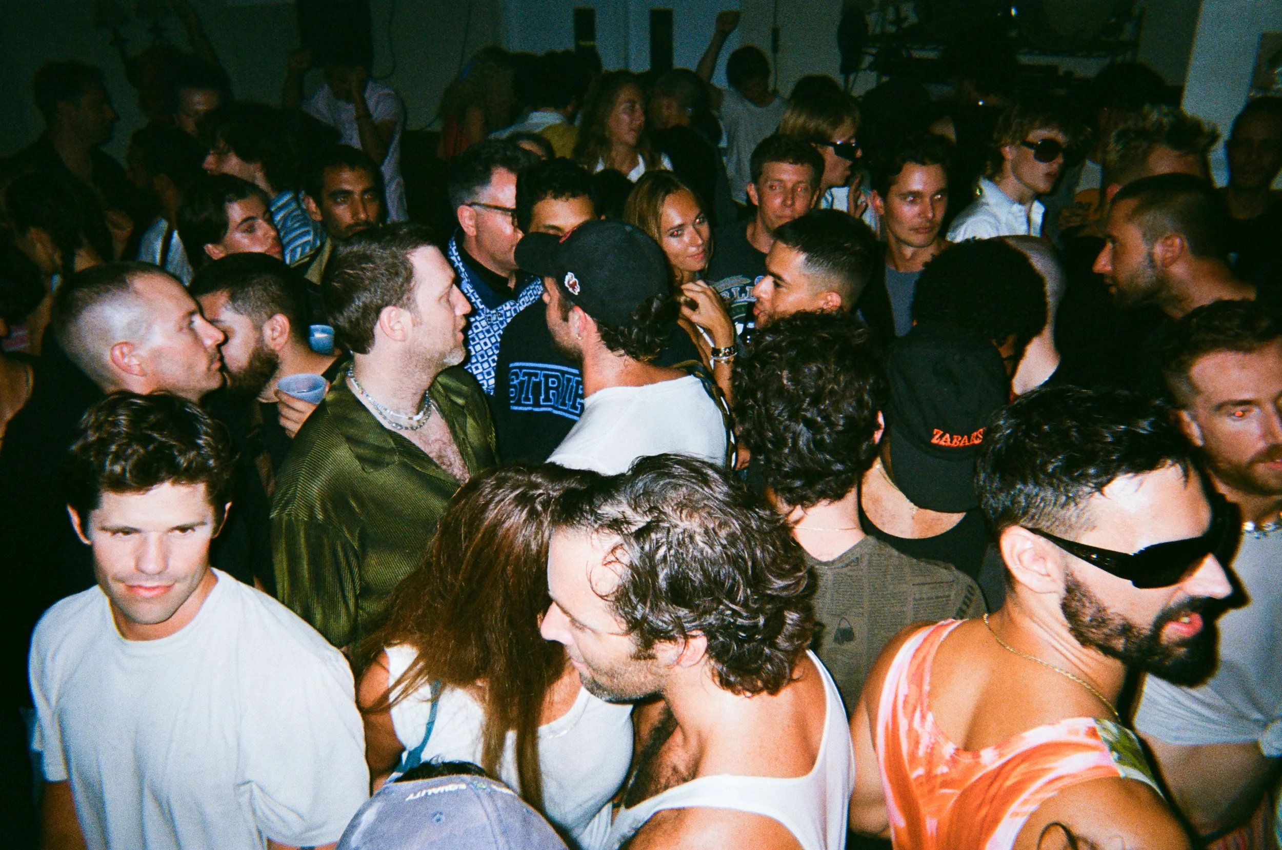 A large crowd of diverse people gathering closely together at an indoor event, some wearing sunglasses, with a dark background.