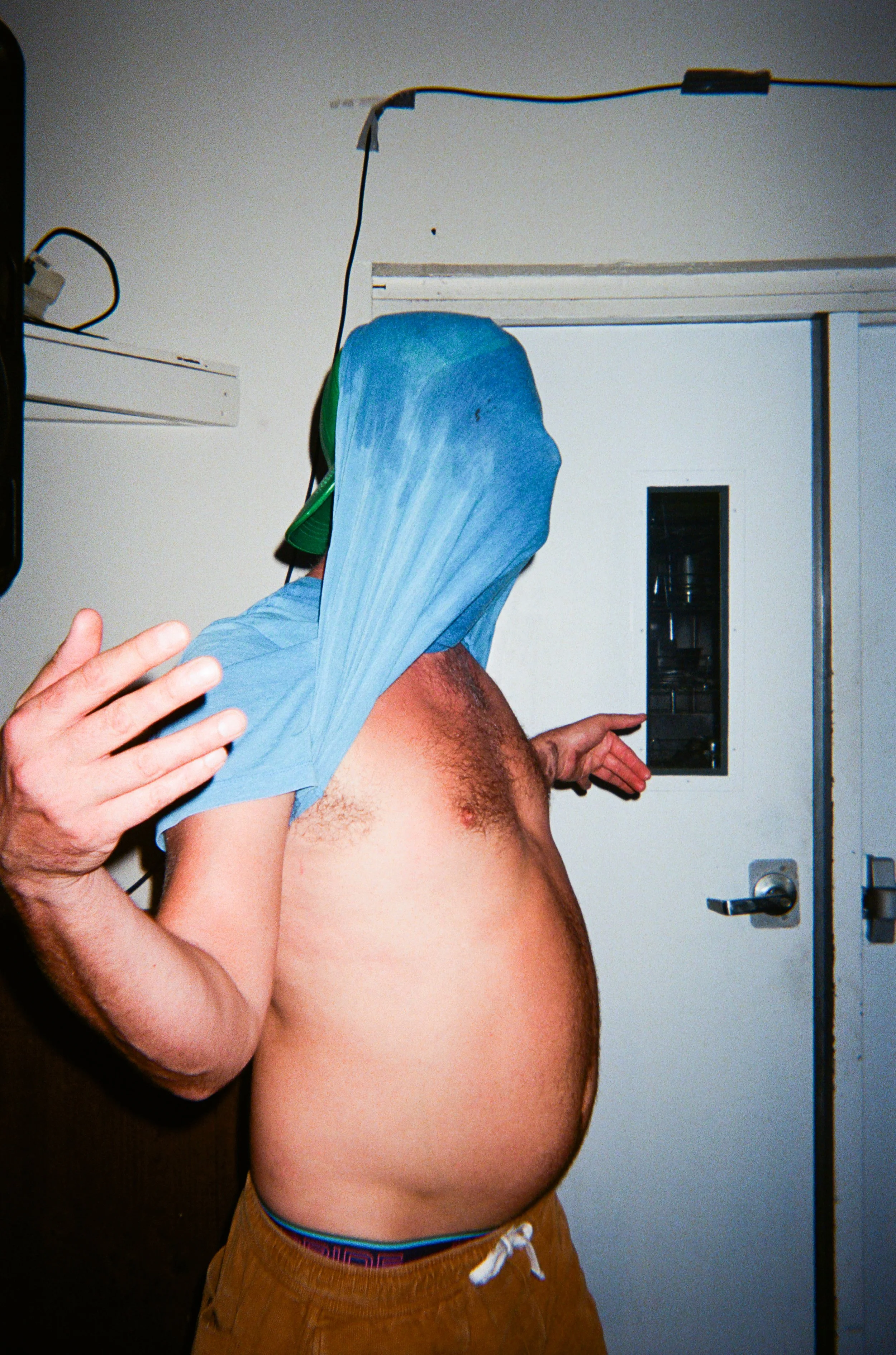 A shirtless man with a large blue plastic bag over his head, standing in front of a white wall with a small window and a door.