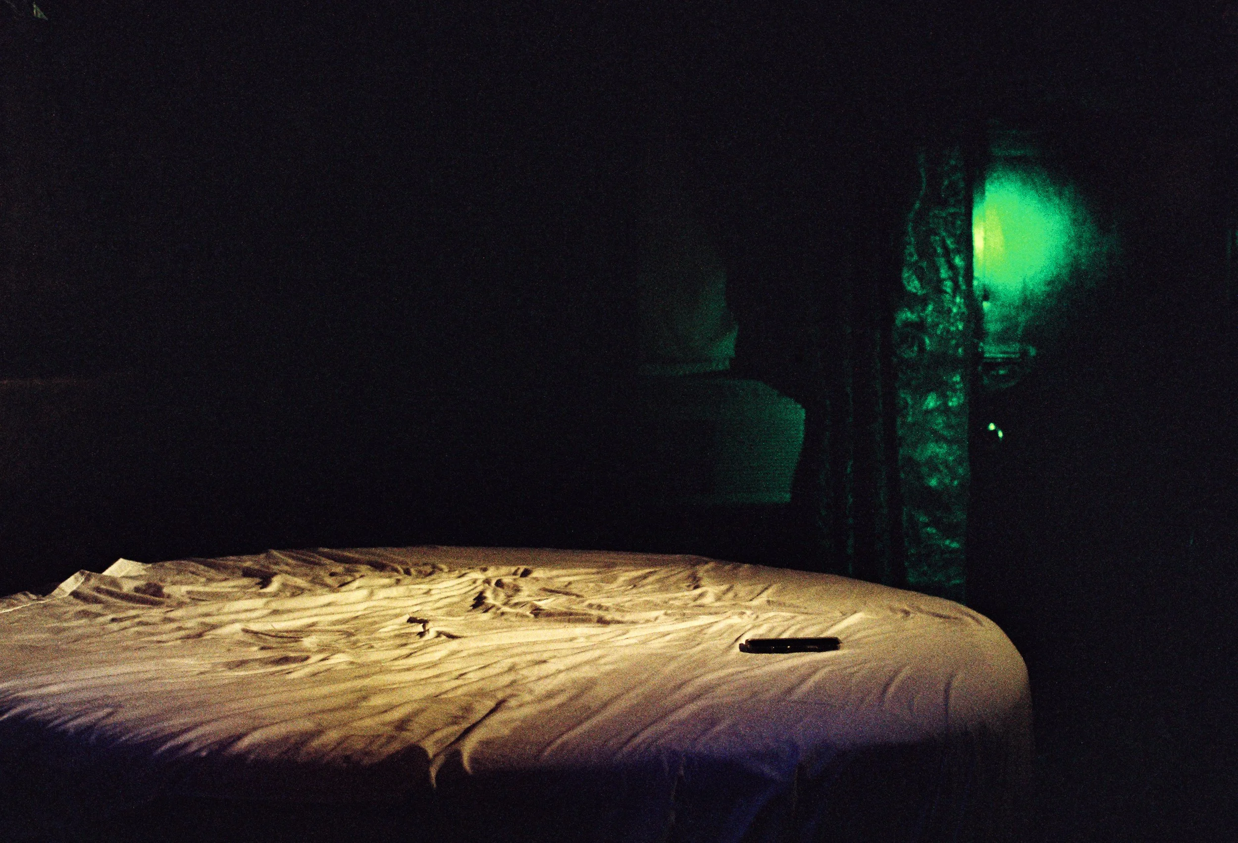 Dark room with a bed that has crumpled bedsheets, and a television or monitor emitting a green glow in the background.