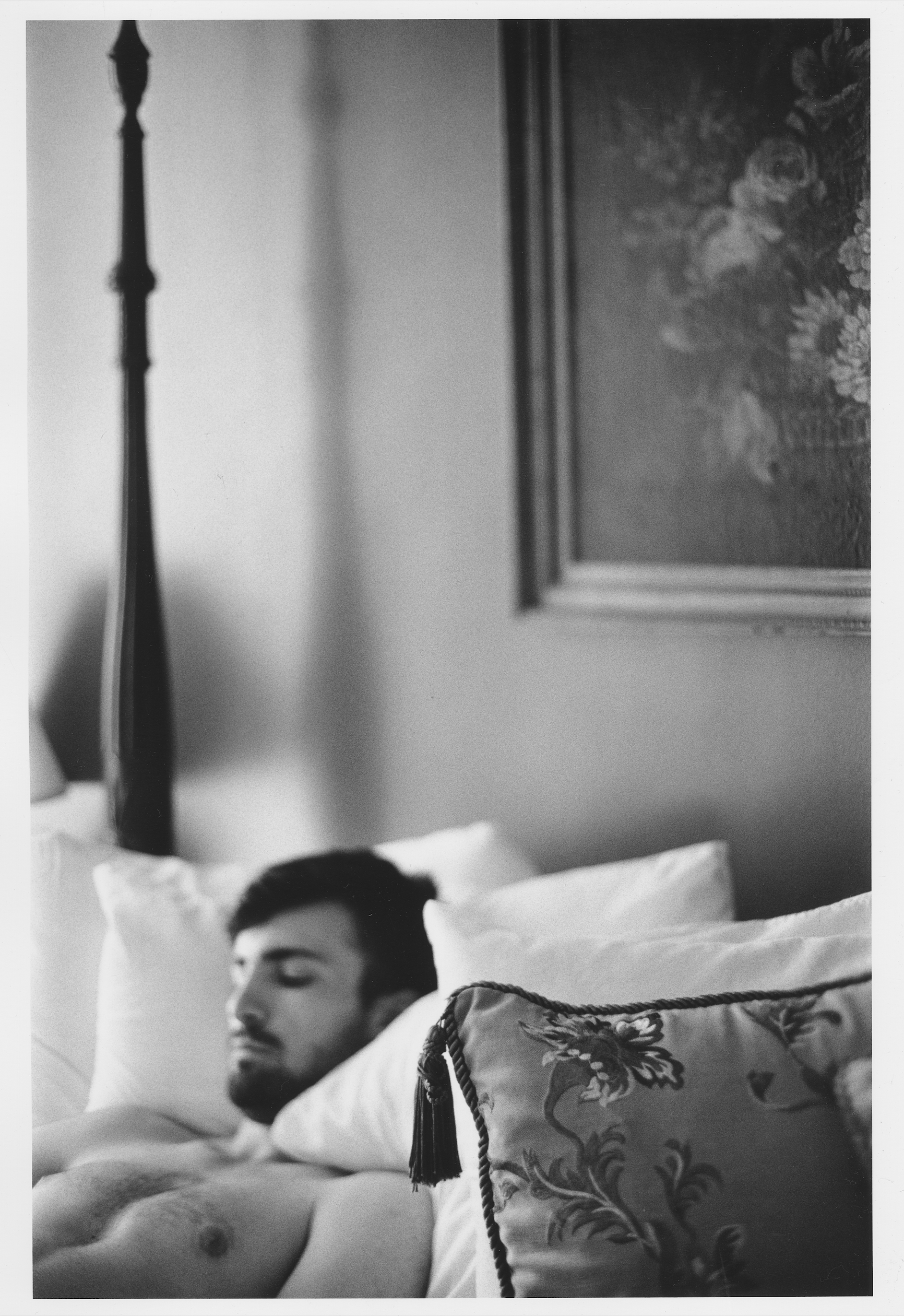 A black and white photo of a young man sleeping in bed with his head resting on a pillow. There is a decorative bag with floral embroidery and tassels on the bed. A framed picture of flowers hangs on the wall in the background.