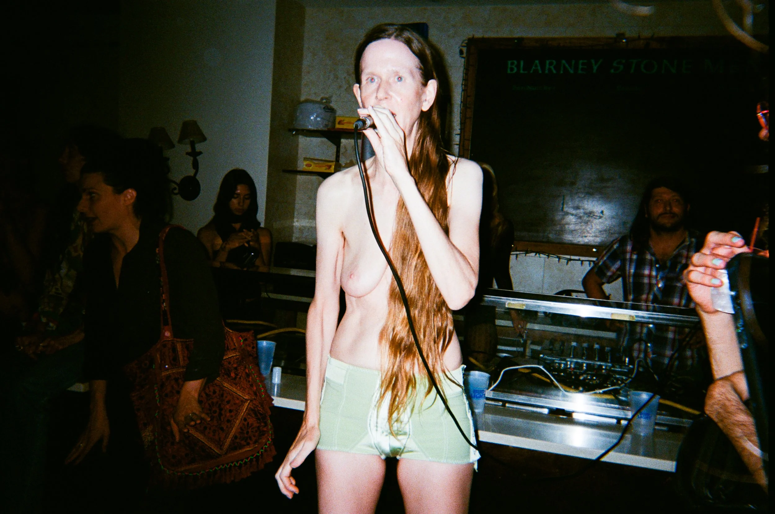 A topless woman with long reddish hair wearing light-colored shorts, holding a microphone, standing in a dimly lit room with multiple people and a blackboard in the background.