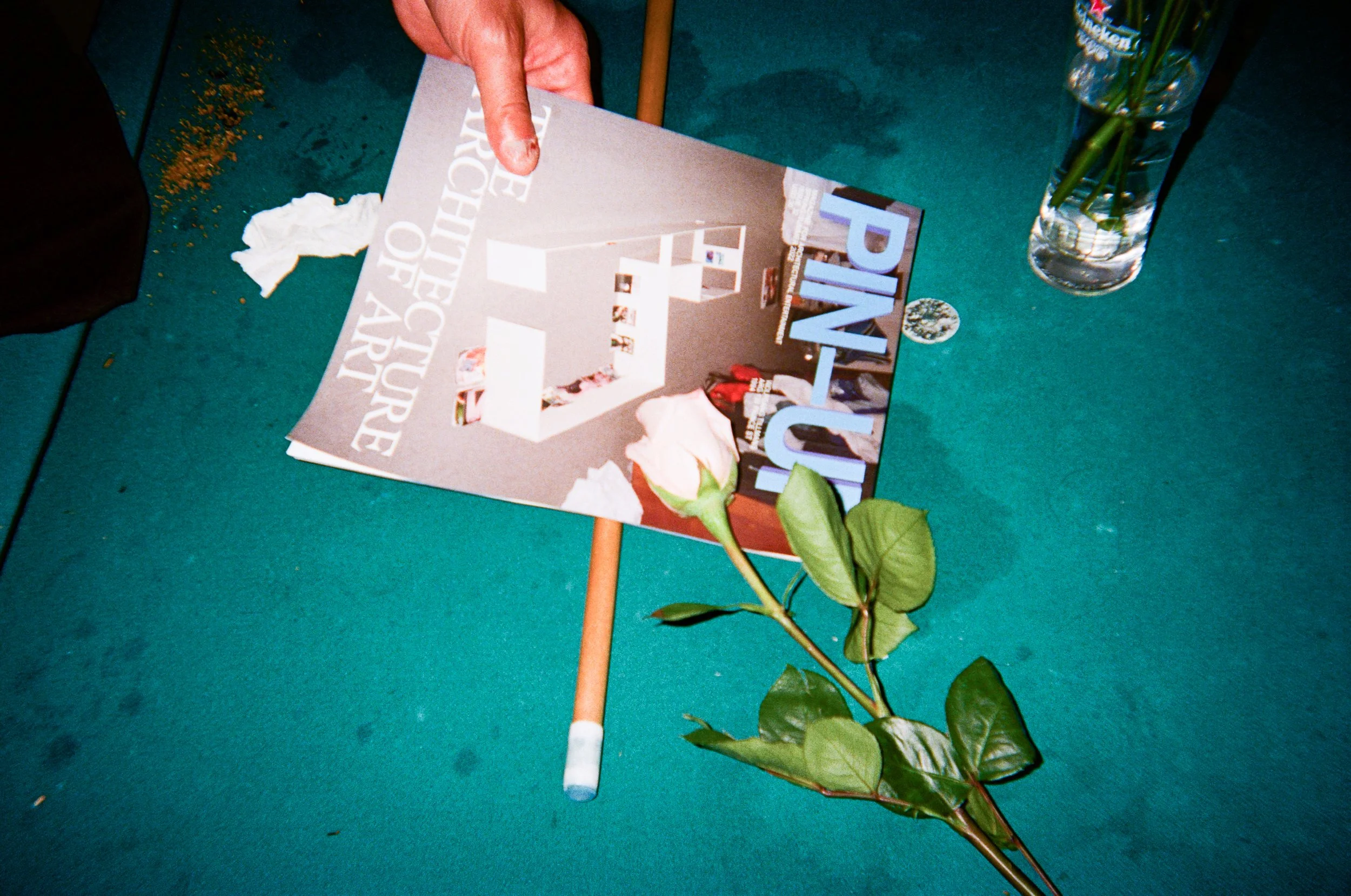A magazine titled 'Pin Up' resting on a green tabletop, partially covered by a rose stem with green leaves. A clear glass vase with water and green stems is also on the table.