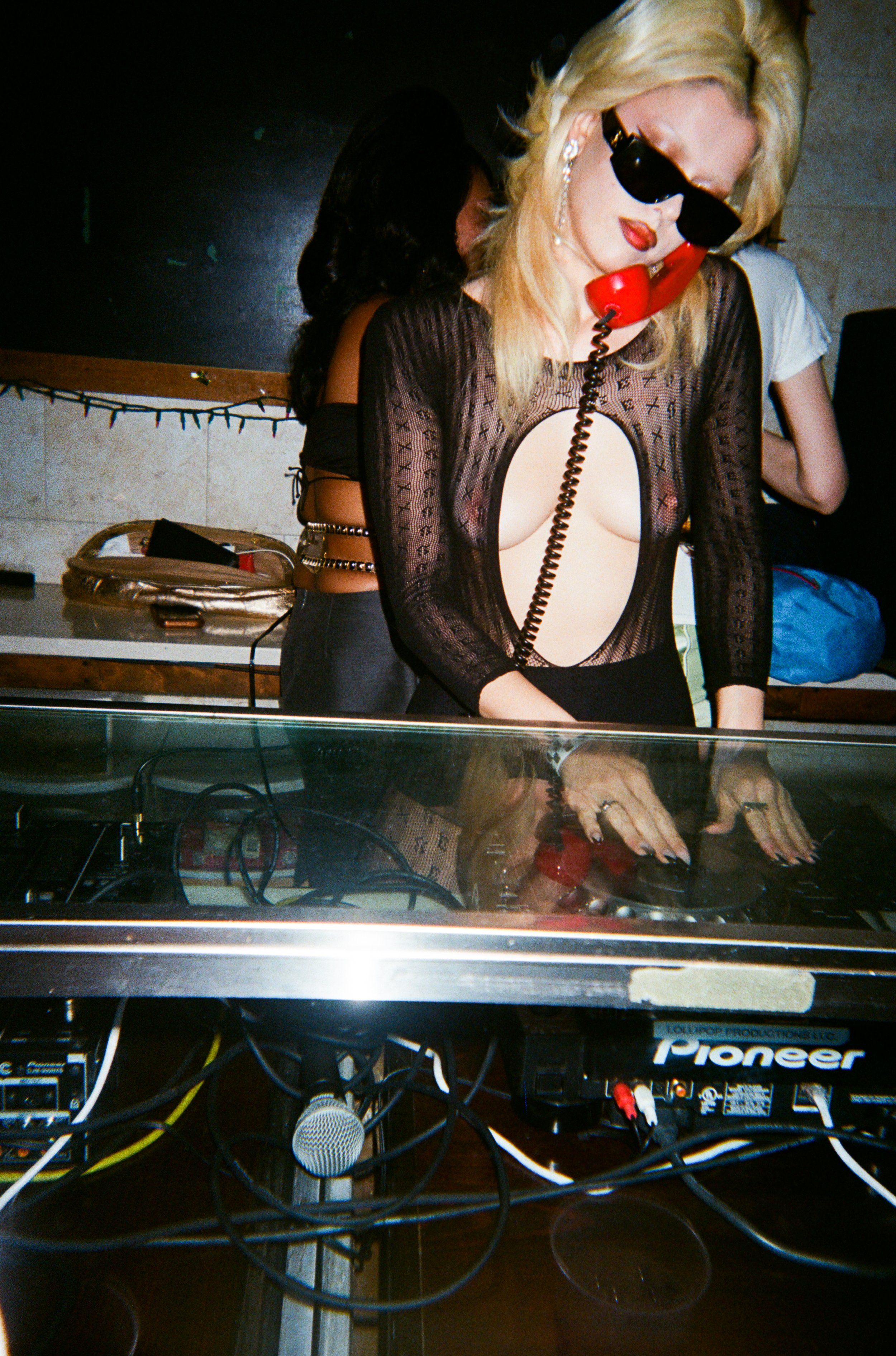 Woman with blonde hair, wearing sunglasses and a sheer black top, speaking into a red telephone at a DJ setup with turntables and audio equipment in a dimly lit room.