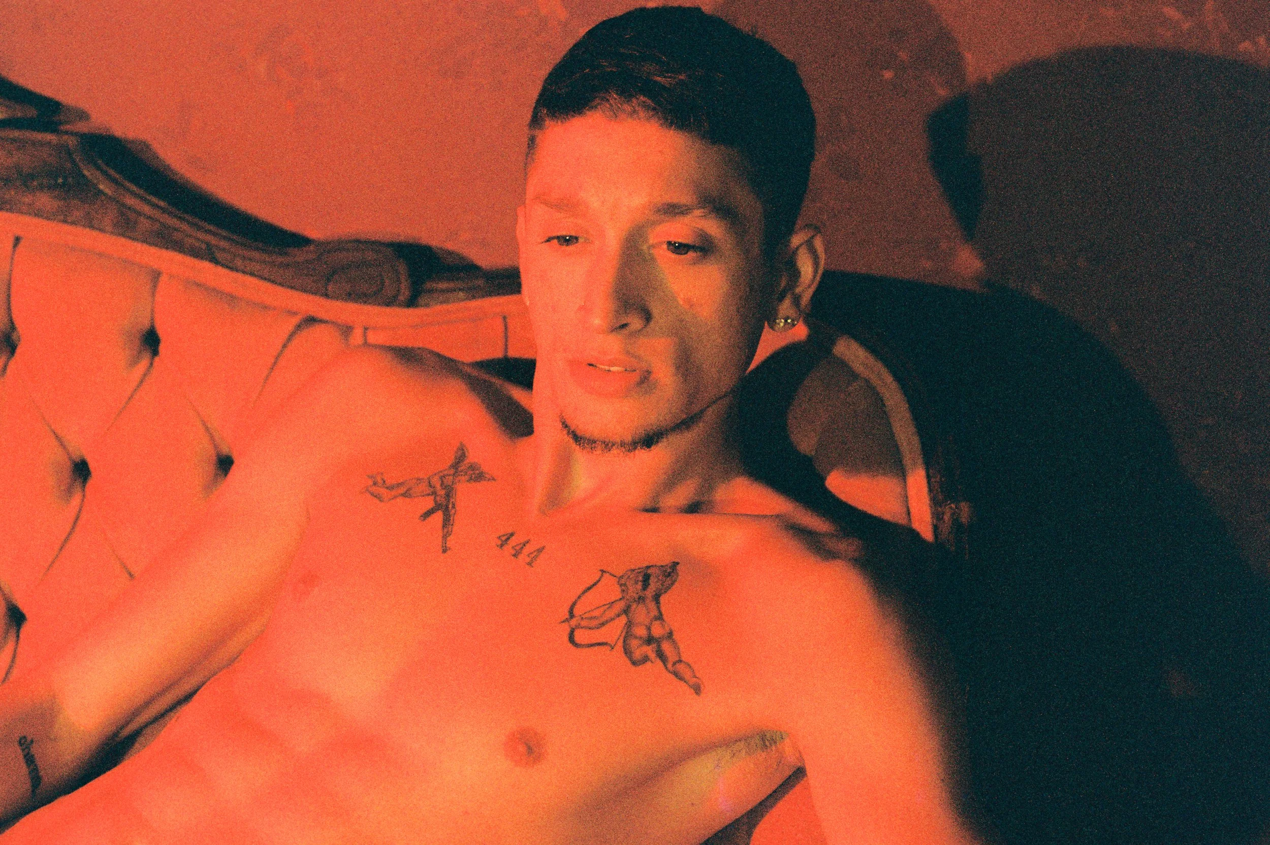 A young man with short dark hair and earrings lying on a bed with a vintage wooden headboard, illuminated by warm red lighting, showcasing tattoos on his shoulder and chest.