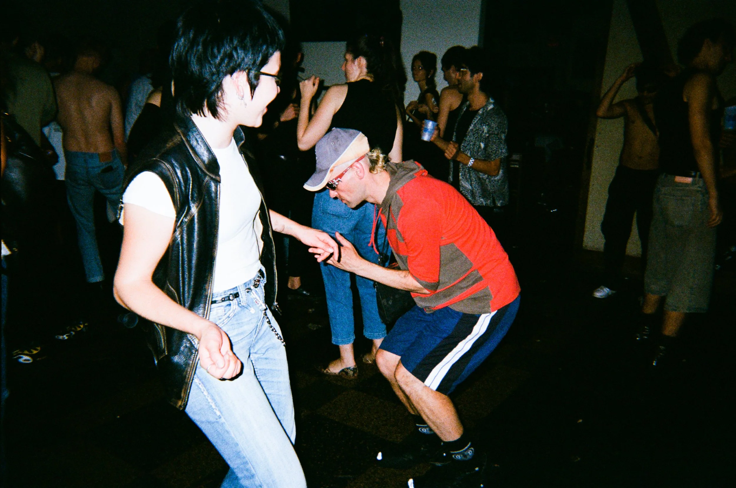 A group of young people dancing and socializing at a party or nightclub in a dimly lit room. The focus is on a young man in a red shirt and cap who is bending over, with others around him talking and enjoying the music.