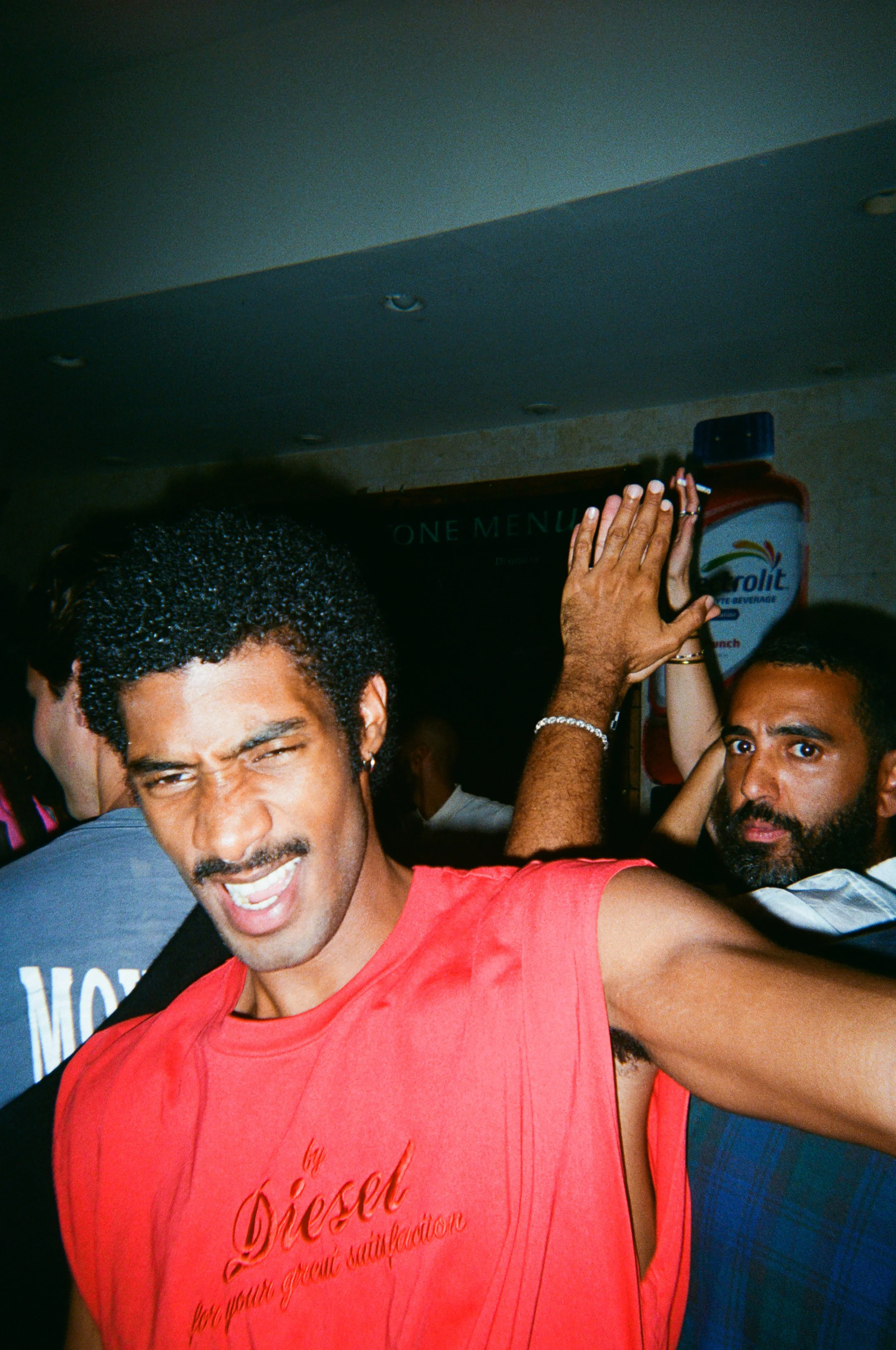 A group of people, mostly men, taking a selfie in a crowded indoor setting. The person in the foreground is wearing a red shirt and making a playful facial expression. There are other people in the background, including a man with a beard wearing a b