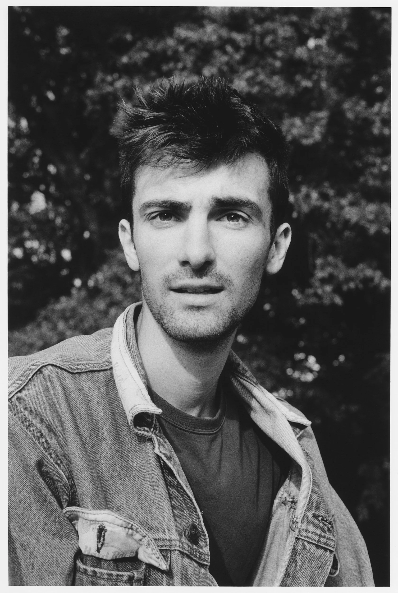 A black-and-white photo of a young man outdoors, wearing a denim jacket over a t-shirt, with short messy hair and a serious expression, blurred trees in the background.