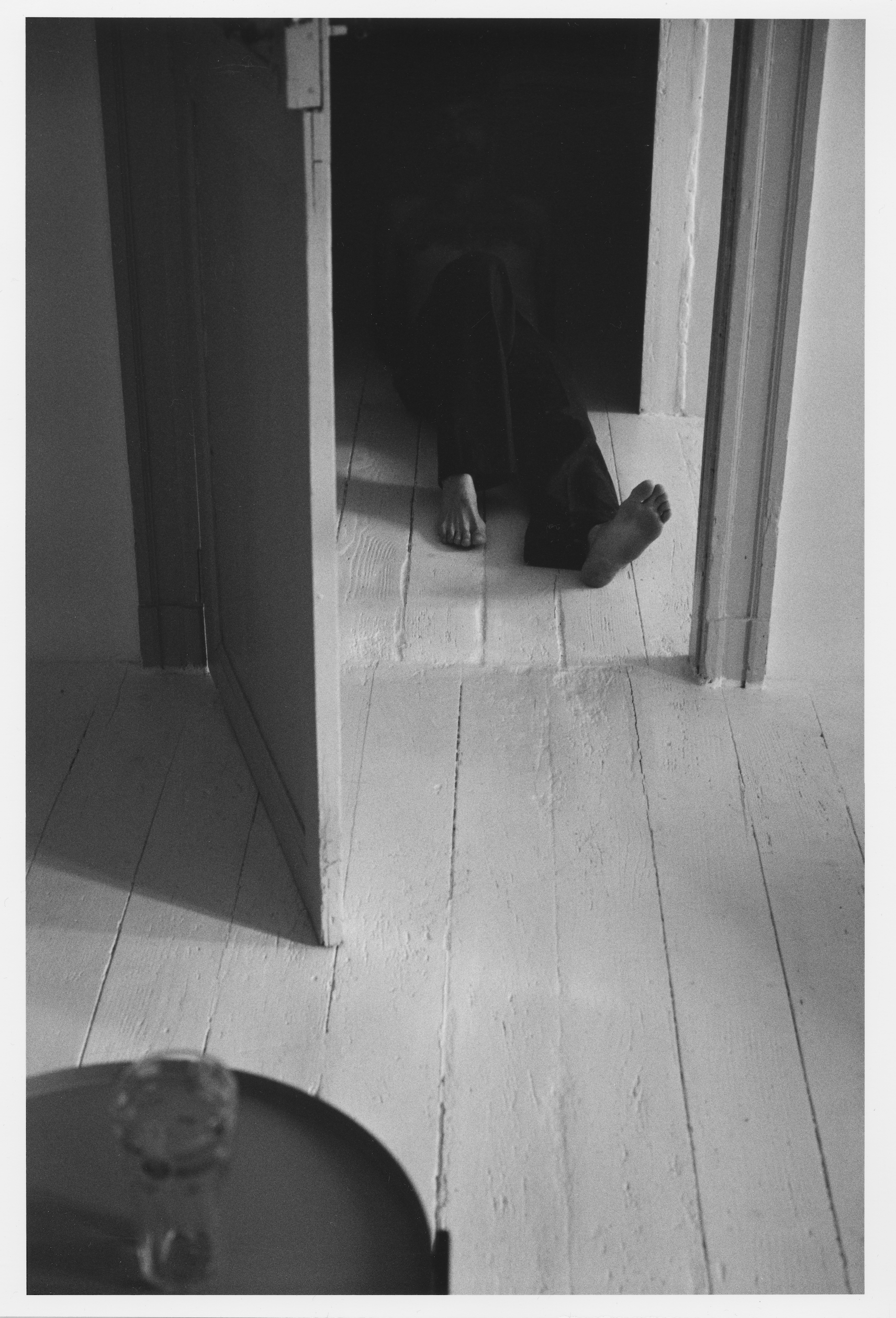 A person lying on their back on the floor with legs extended out, viewed through a mirror. The person appears to be taking a photo of their feet with a camera, with a dark background behind them. The perspective is from an angle near the person's hea