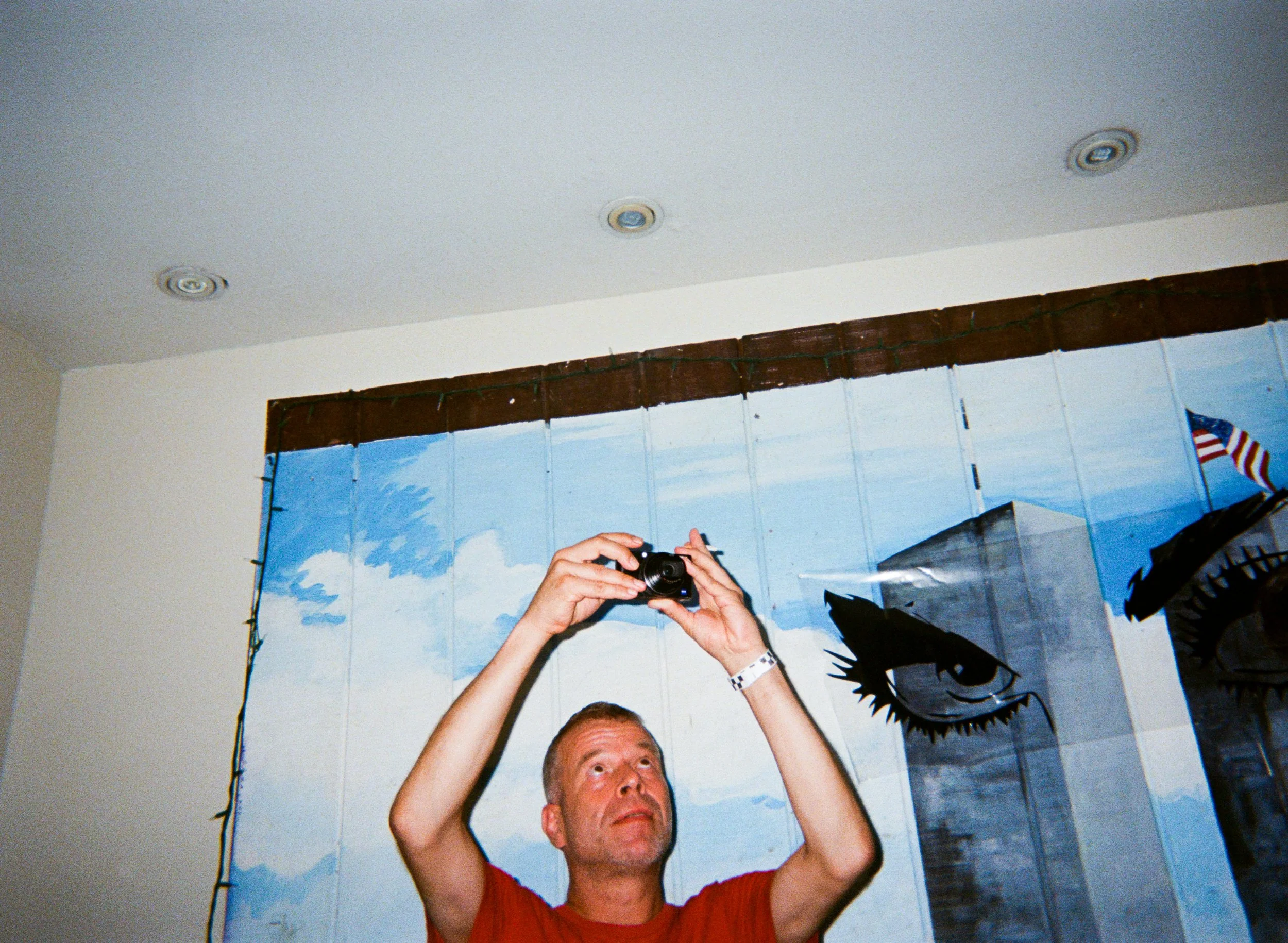 Person holding a camera, partially visible head at the bottom, in front of a large mural of a blue sky with clouds, and a portrait of a shark with an open mouth on the right.