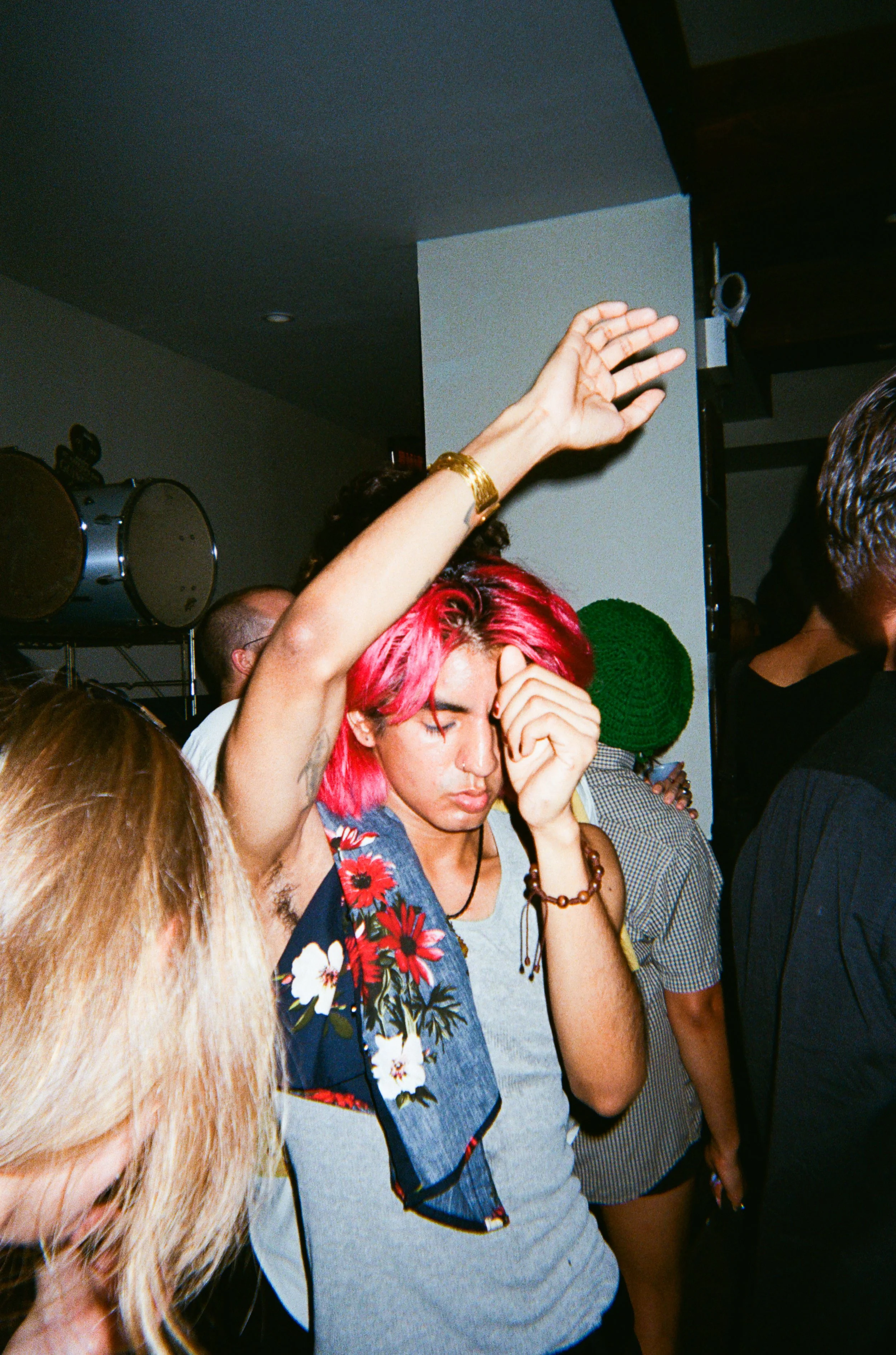 A person with bright pink hair is at a crowded indoor event, touching their forehead with one hand, wearing a gray shirt with a floral and plaid design scarf, a brown bracelet, and a necklace. Other people are in the background, some with colorful ha
