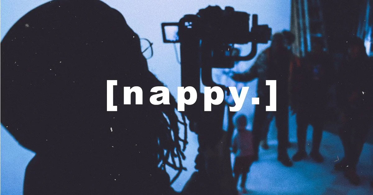 Silhouette of a person operating a camera, with a group of people and children in the background in an indoor setting, overlaid with the word '[nappy.]'