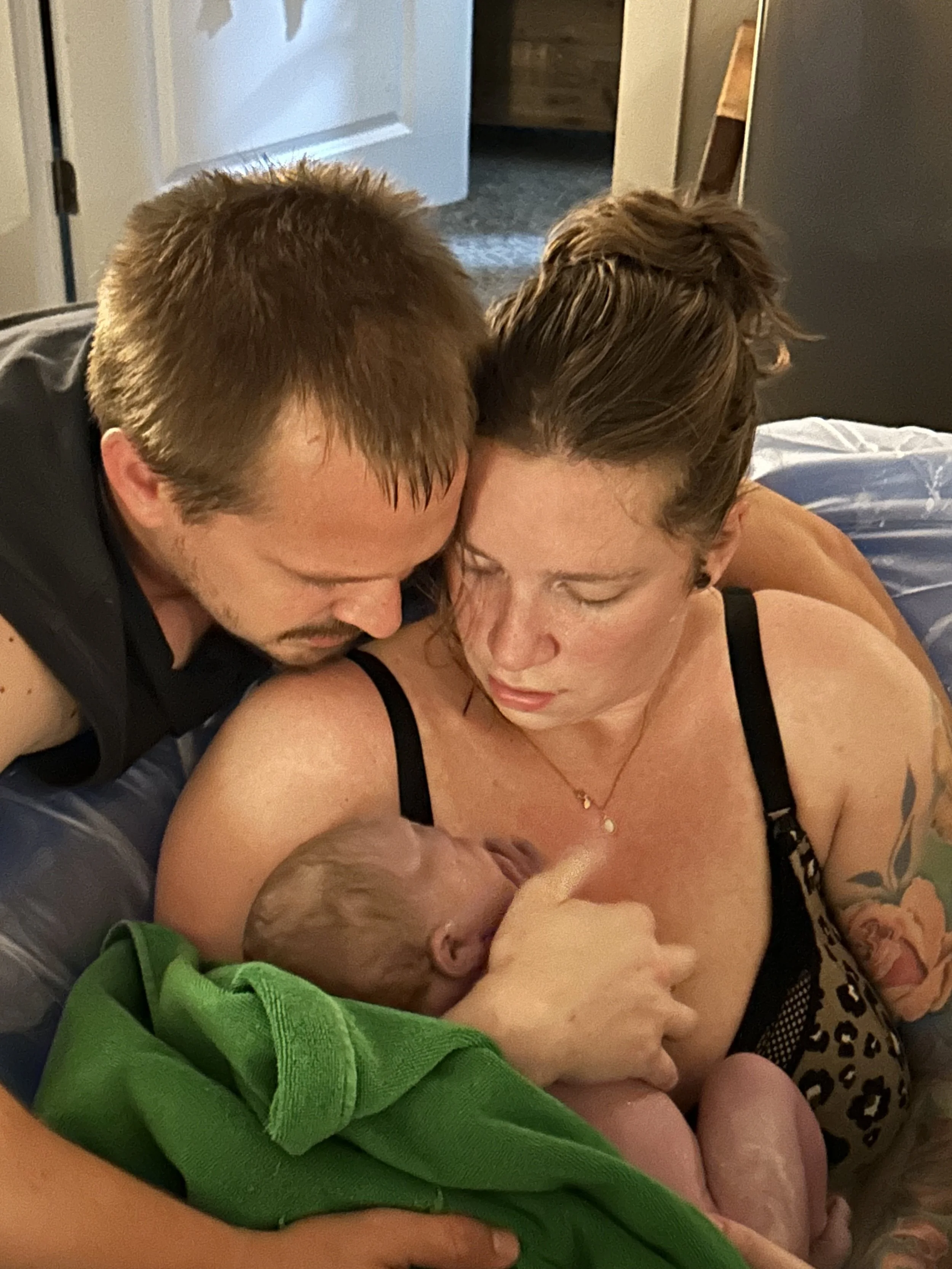A woman with tattoos on her arm, wearing a black bra, breastfeeding a newborn baby. A man leaning over from the side, also looking at the baby, with a short beard and mustache, in a dark shirt. They are in a cozy indoor setting, possibly a bedroom.
