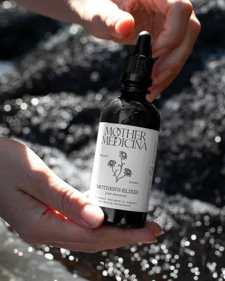 Hands holding a dark glass dropper bottle labeled "Mother Medica Mother's Elixir with chamomile," with a blurred natural background.