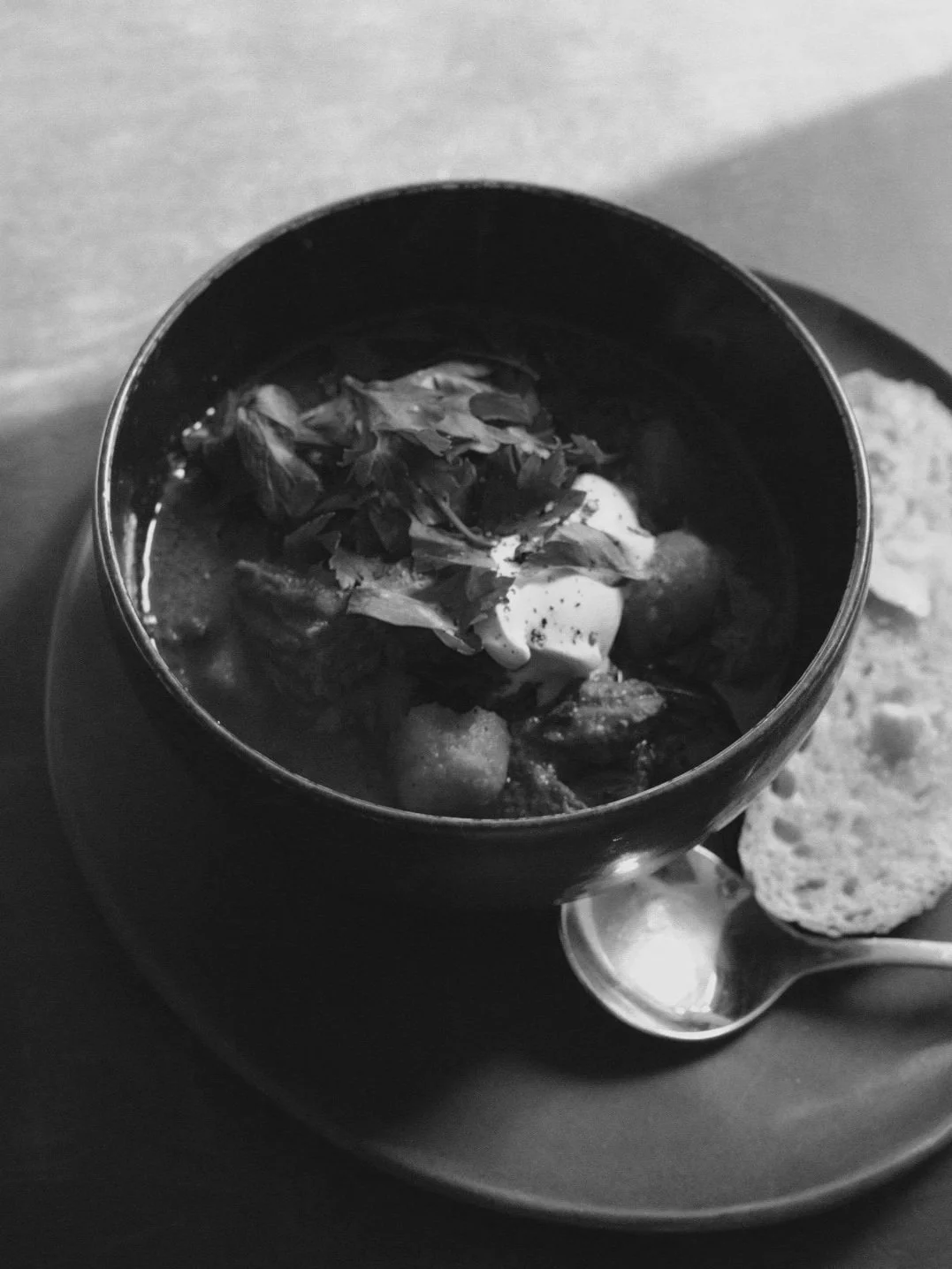 Black and white photo of a bowl of soup with leafy herbs and what appears to be a dollop of cream. A slice of bread and a spoon are on the plate beside it.