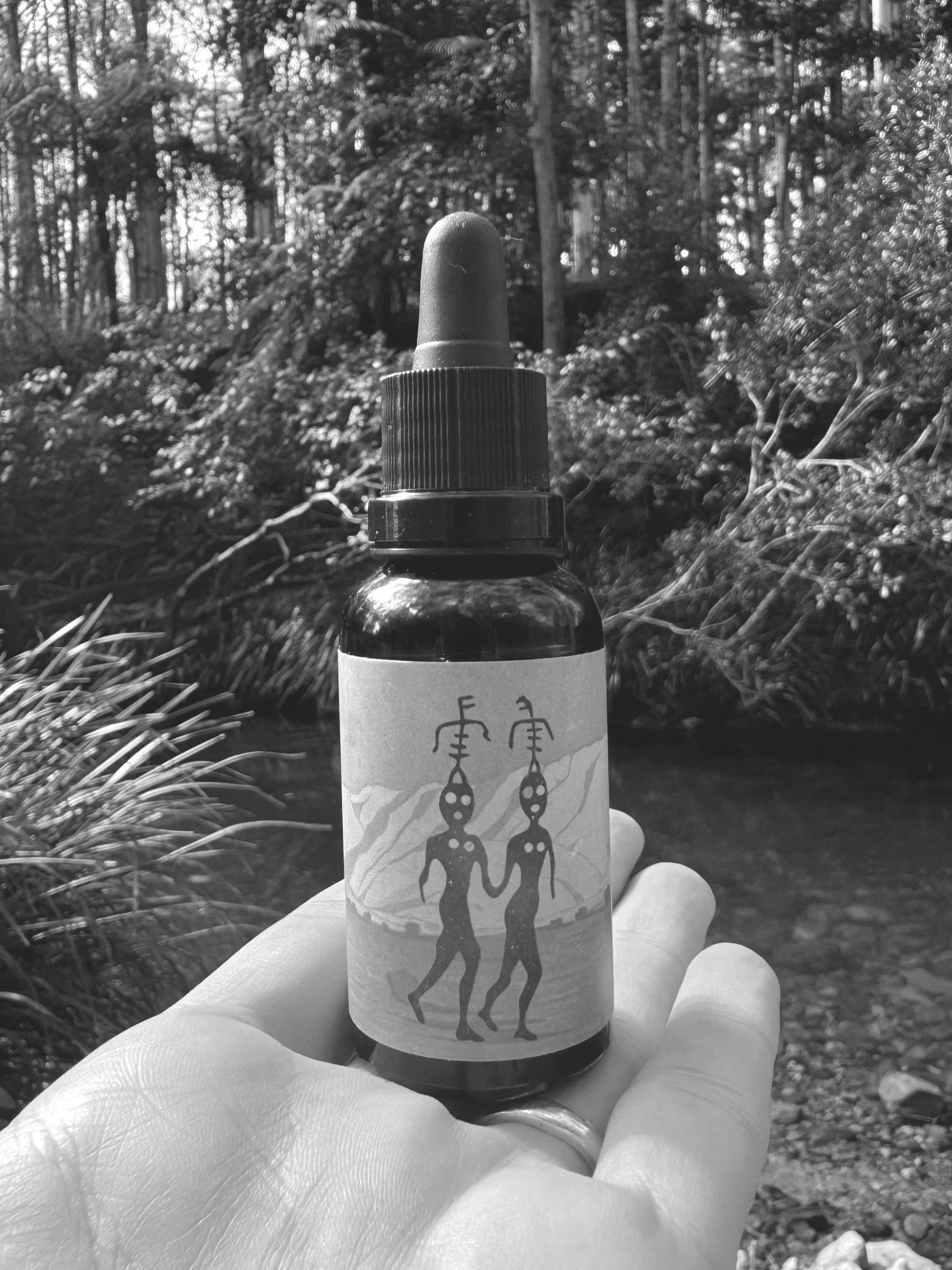 Hand holding a small dropper bottle with an abstract figure label, forest background