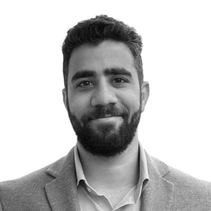 A man with a beard and curly hair smiling in a suit and collared shirt against a plain white background.