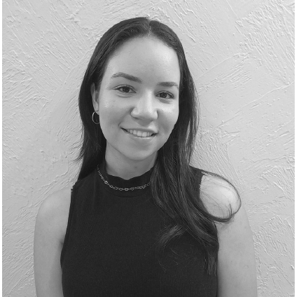 A young woman with long dark hair, smiling, wearing a sleeveless black top and a necklace, standing against a textured light-colored wall.