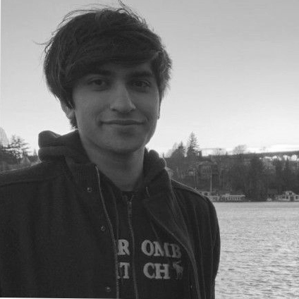 A young man with dark, tousled hair, wearing a hoodie and jacket, smiling outdoors near a body of water with trees and buildings in the background.
