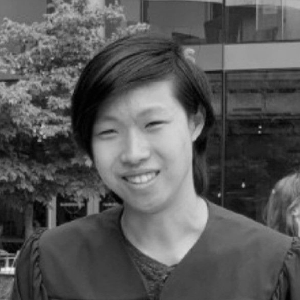 A young woman with short dark hair smiling outdoors, wearing a graduation gown.