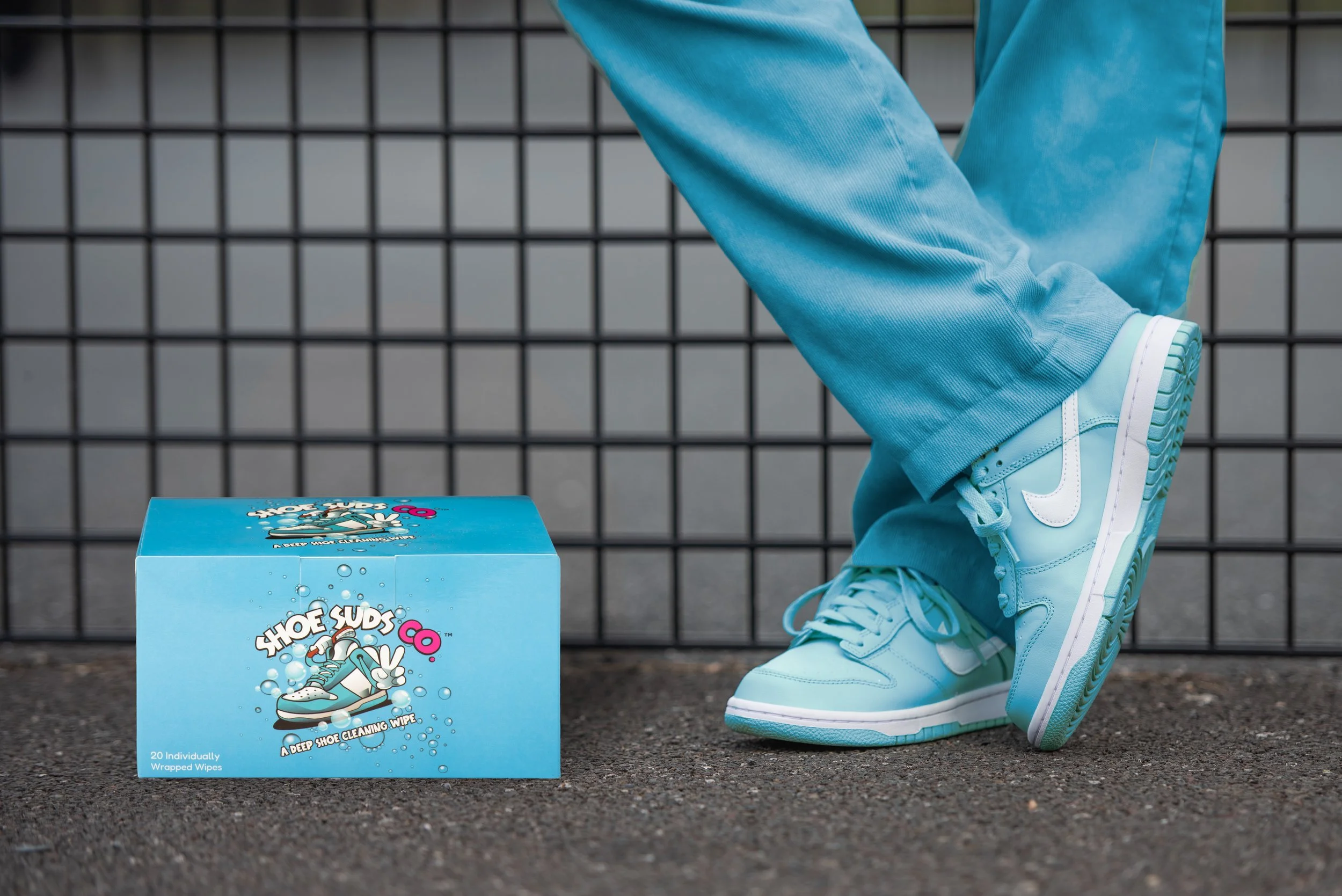 Close-up of feet in turquoise sneakers with white Nike swoosh, next to a blue box of Shoe Suds shoe cleaning wipes on the ground, with a black wire fence in the background.