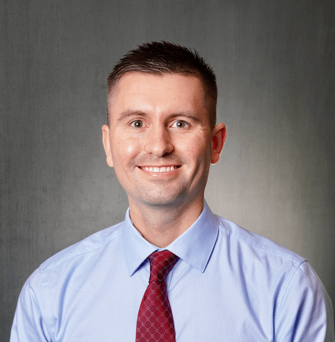 Man in a blue shirt and red tie smiling against a gray background.