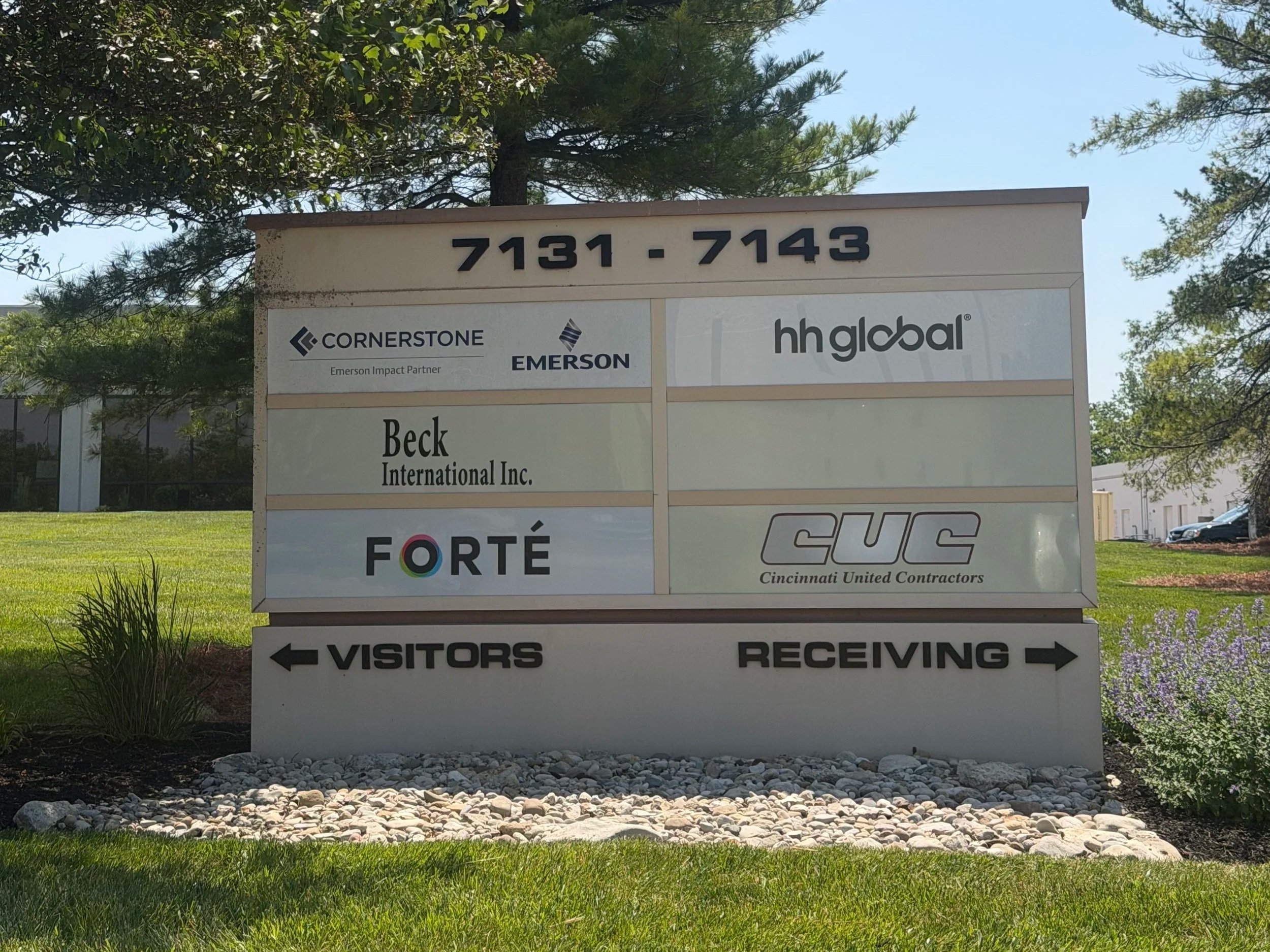 A signboard outdoors with multiple company logos and contact information at the top. The sign has arrows pointing left and right indicating directions for visitors and receiving areas. The background includes trees, a clear sky, and a grassy area.