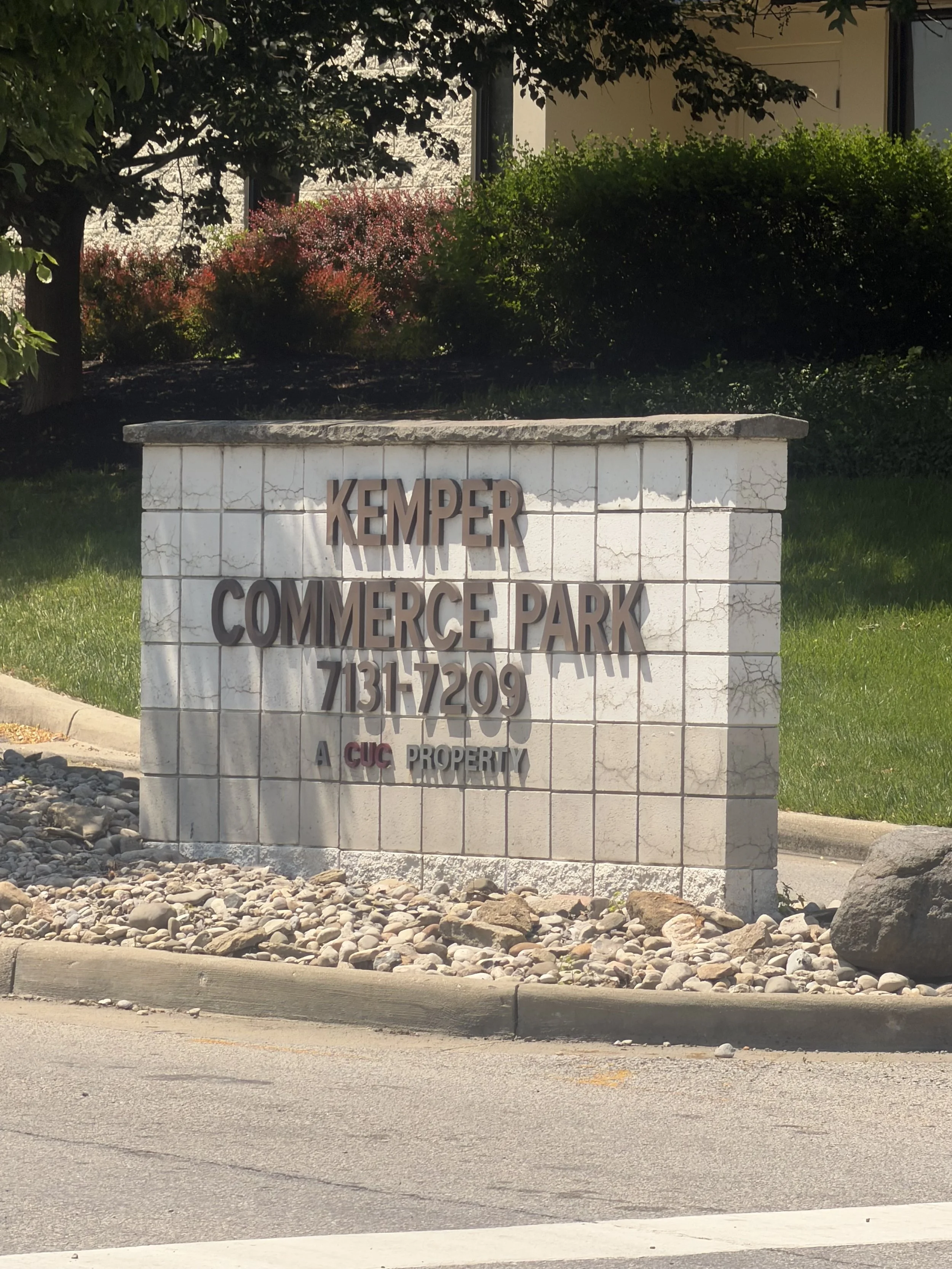 A white stone sign with the words "Kemper Commerce Park" in large letters, situated on a bed of rocks with grass and trees in the background.