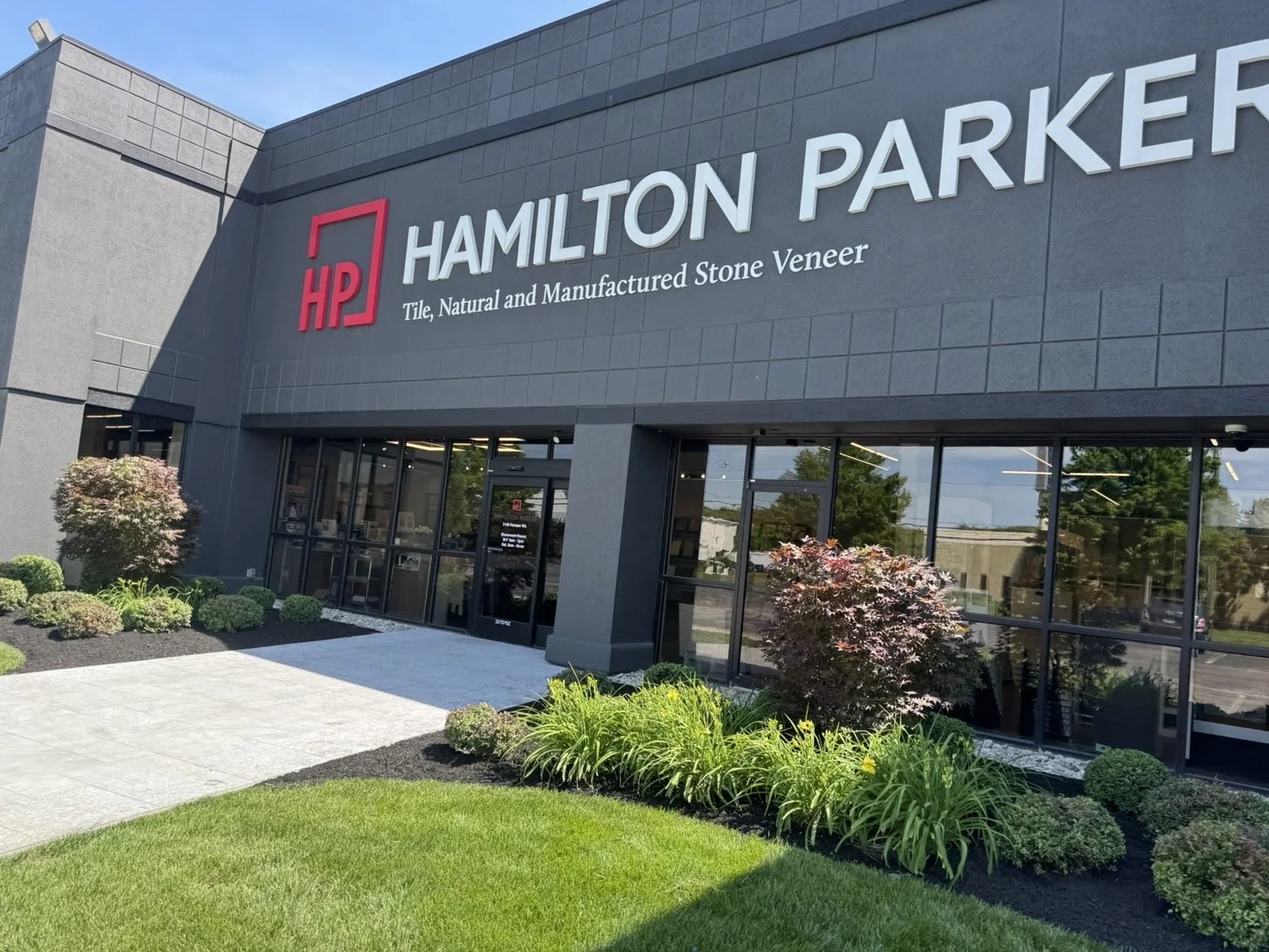 Front view of a modern commercial building with a sign that reads \"Hamilton Parker\", with the logo \"HP\" in red and the slogan \"Tile, Natural and Manufactured Stone Veneer\". There are landscaped bushes and flowers in front of the building and a 