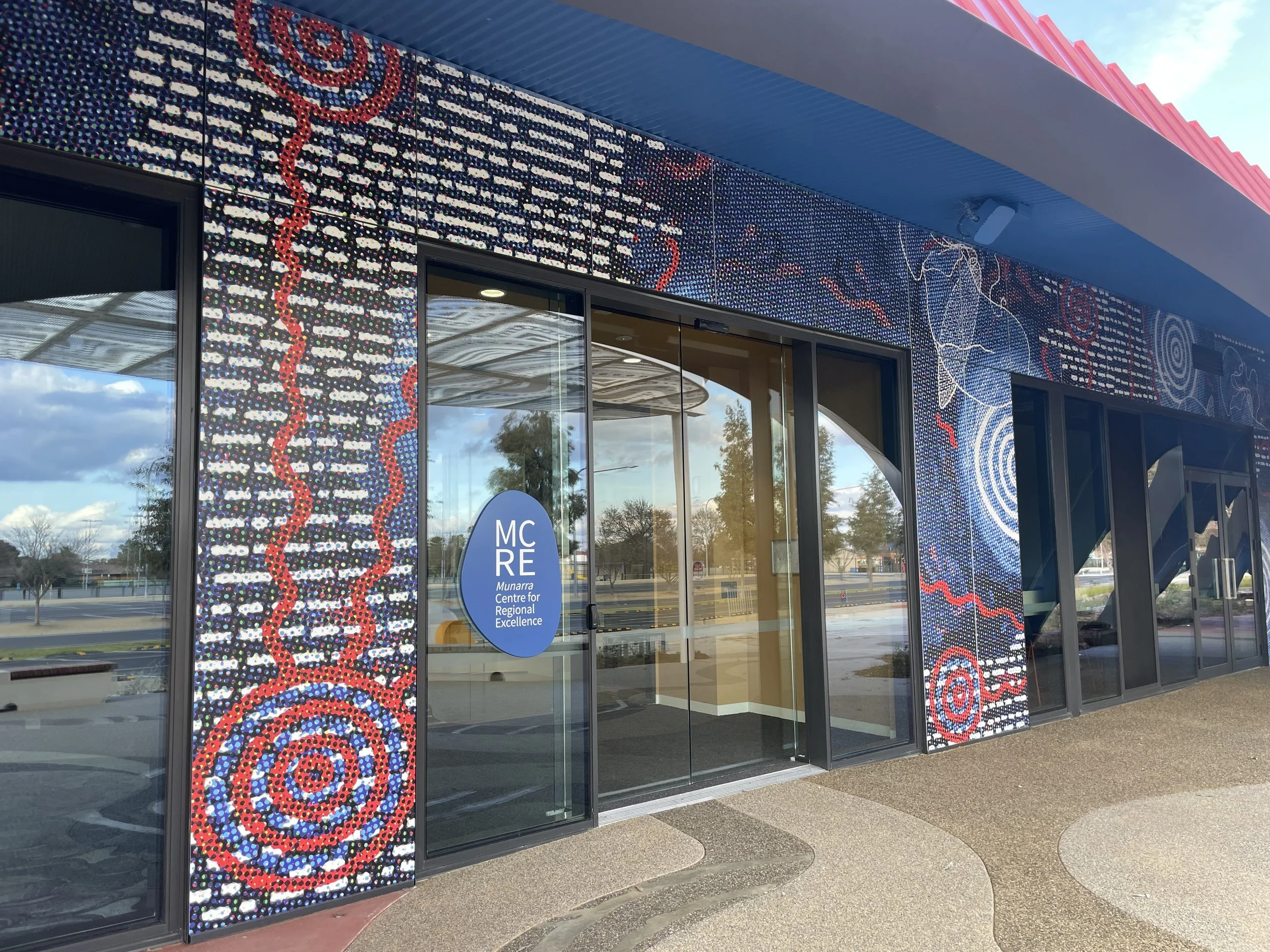 Exterior of the Munarra Centre for Regional Excellence with a colorful, Aboriginal-style dot painting mural on the facade, reflecting the sky and surroundings in the glass doors.