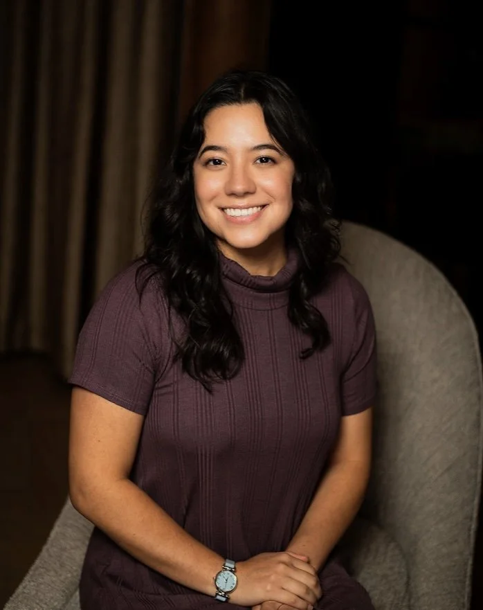 A woman with dark, wavy hair and a bright smile, wearing a purple short-sleeve top, sitting on a beige armchair in a warmly lit room.