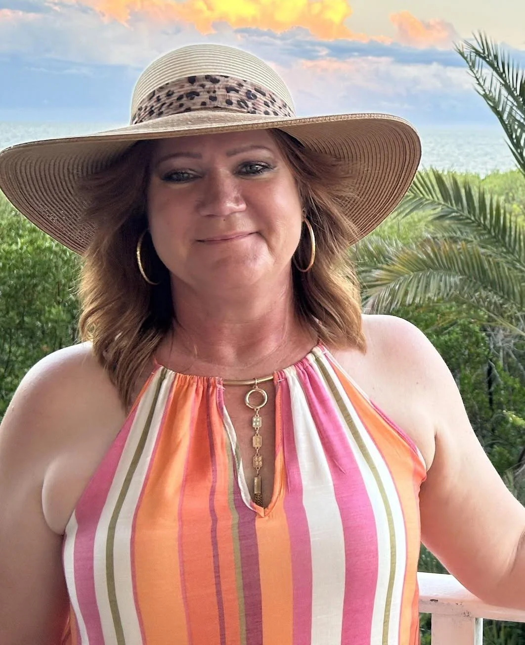 Woman wearing a large straw hat with leopard print band, colorful striped top, gold hoop earrings, and gold jewelry, standing outdoors with greenery, palm trees, ocean, and sunset sky in the background.