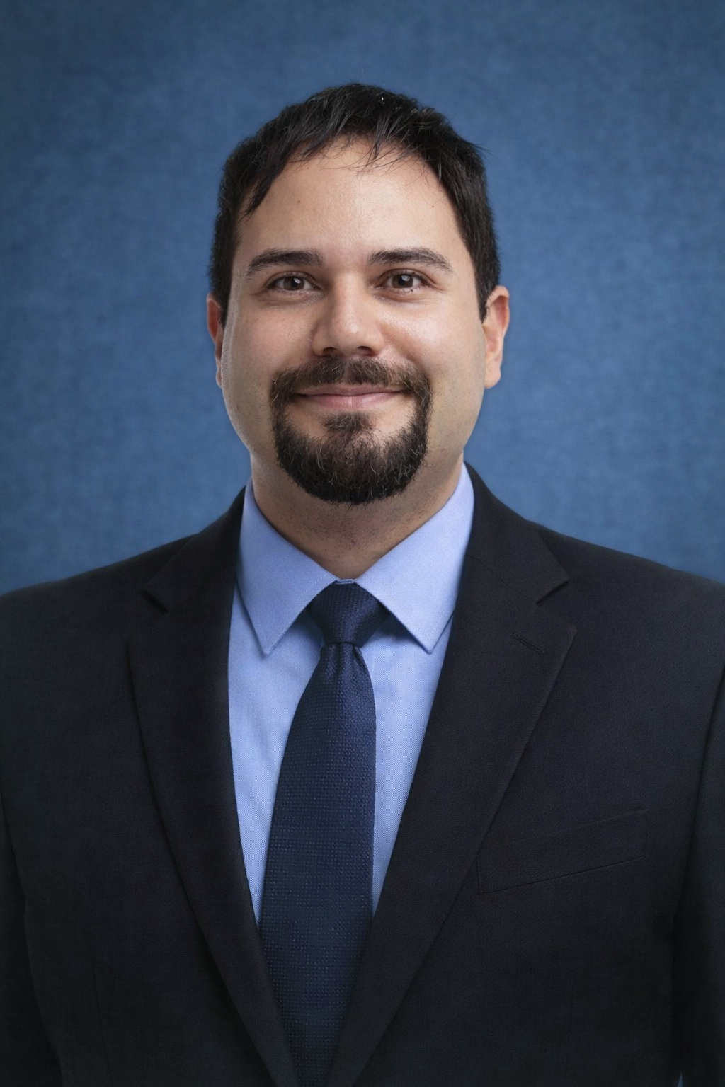 Professional headshot of a man with dark hair, beard, mustache, wearing a dark suit, light blue shirt, and dark tie, smiling in front of a blue background.