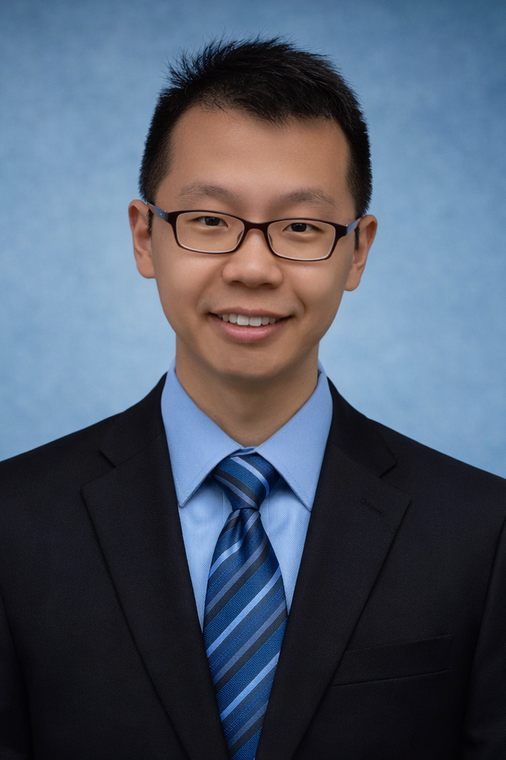 Professional headshot of a young man wearing glasses, a dark suit, light blue shirt, and striped blue tie, smiling against a blue background.