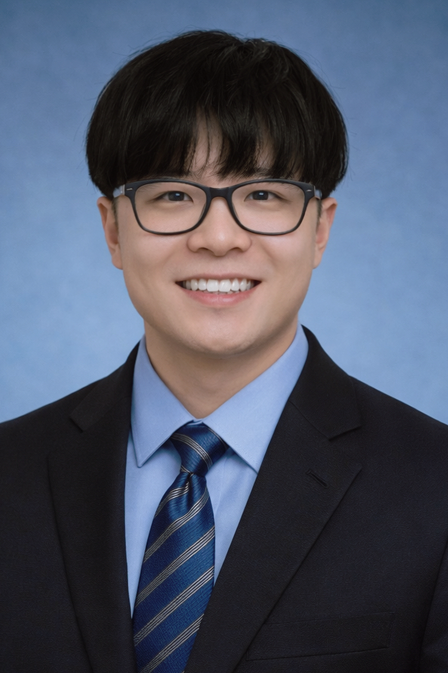 Young man with black hair and glasses, wearing a navy suit, light blue shirt, and striped blue tie, smiling against a blue background.