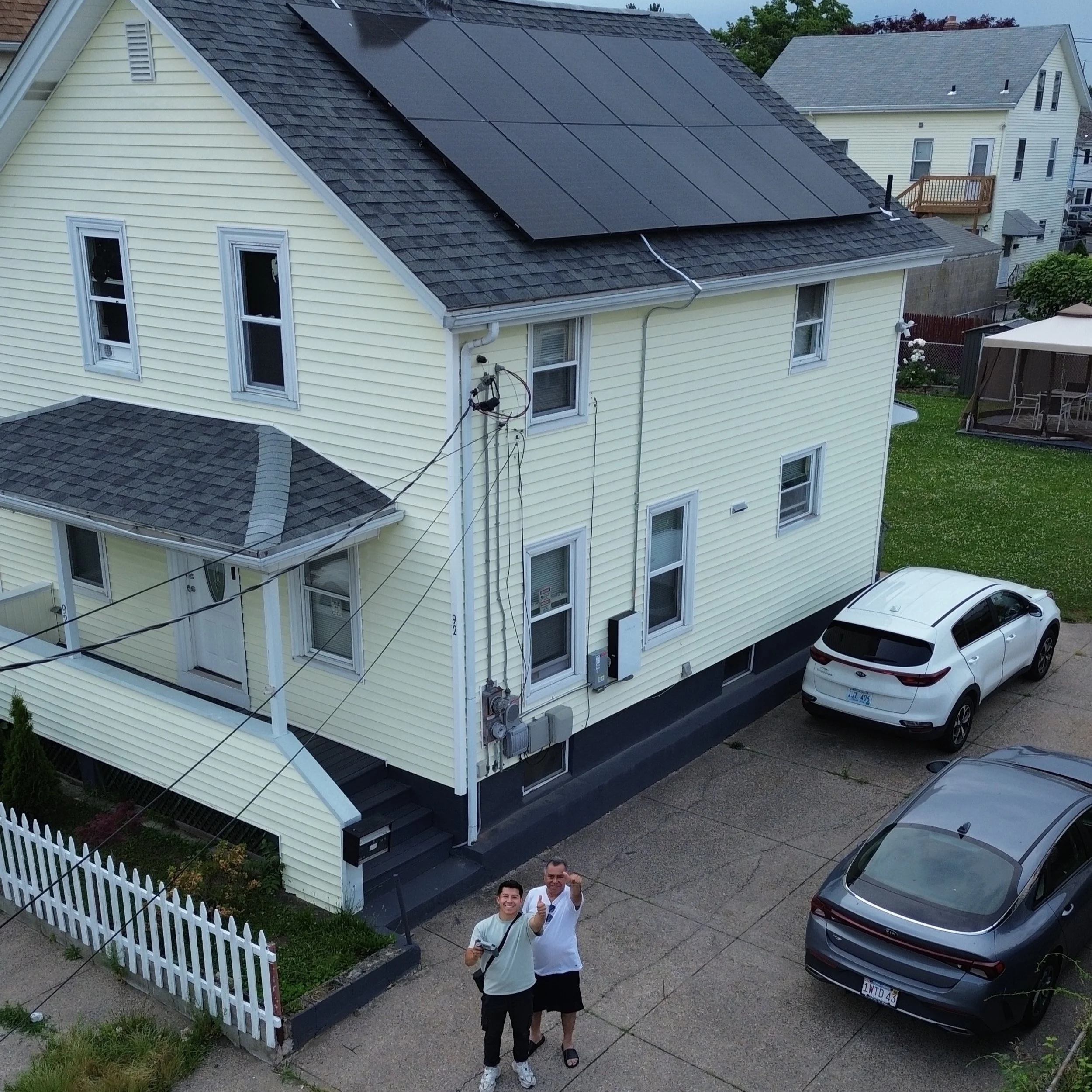 Residential house with solar panels on roof, two cars parked in driveway, two people standing outside pointing up, white picket fence, overcast weather.