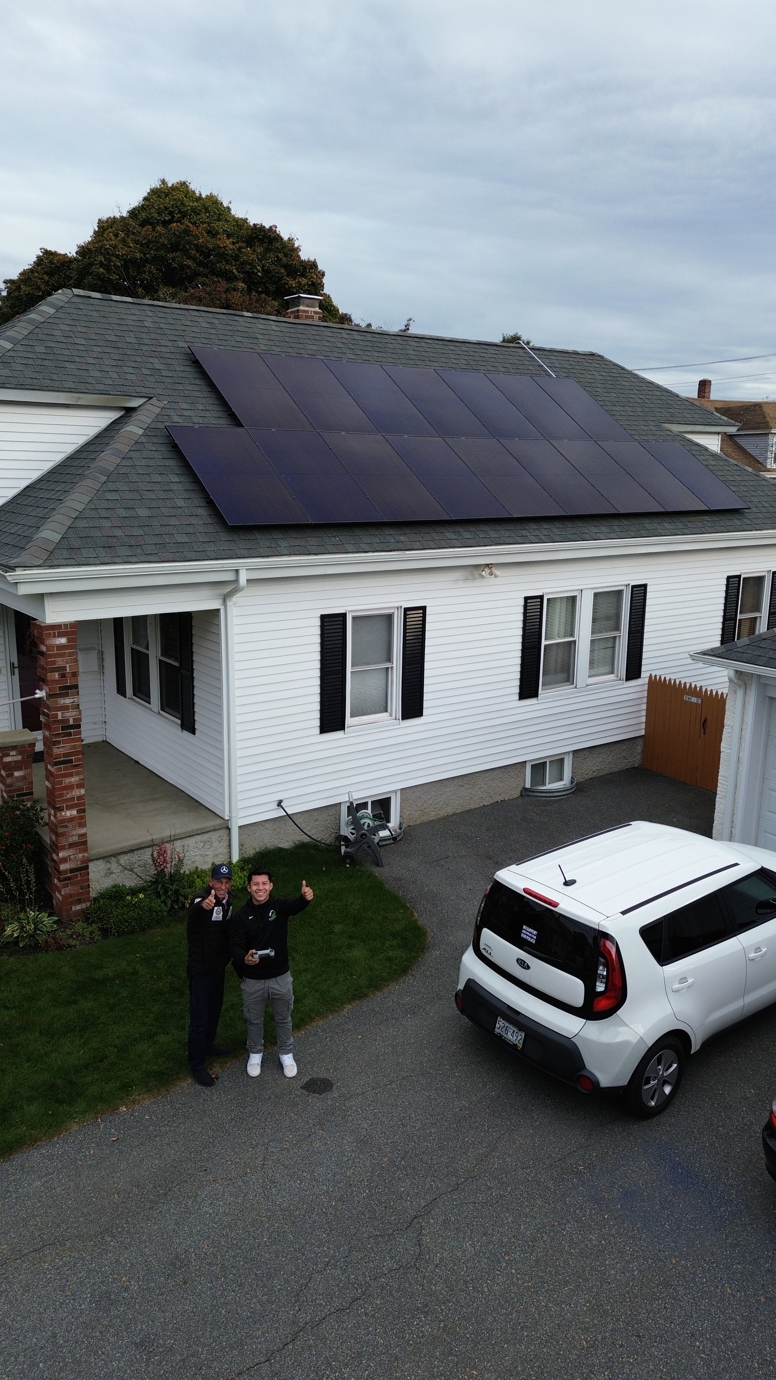 House with solar panels on the roof, two people standing on the driveway, and a white car parked nearby.