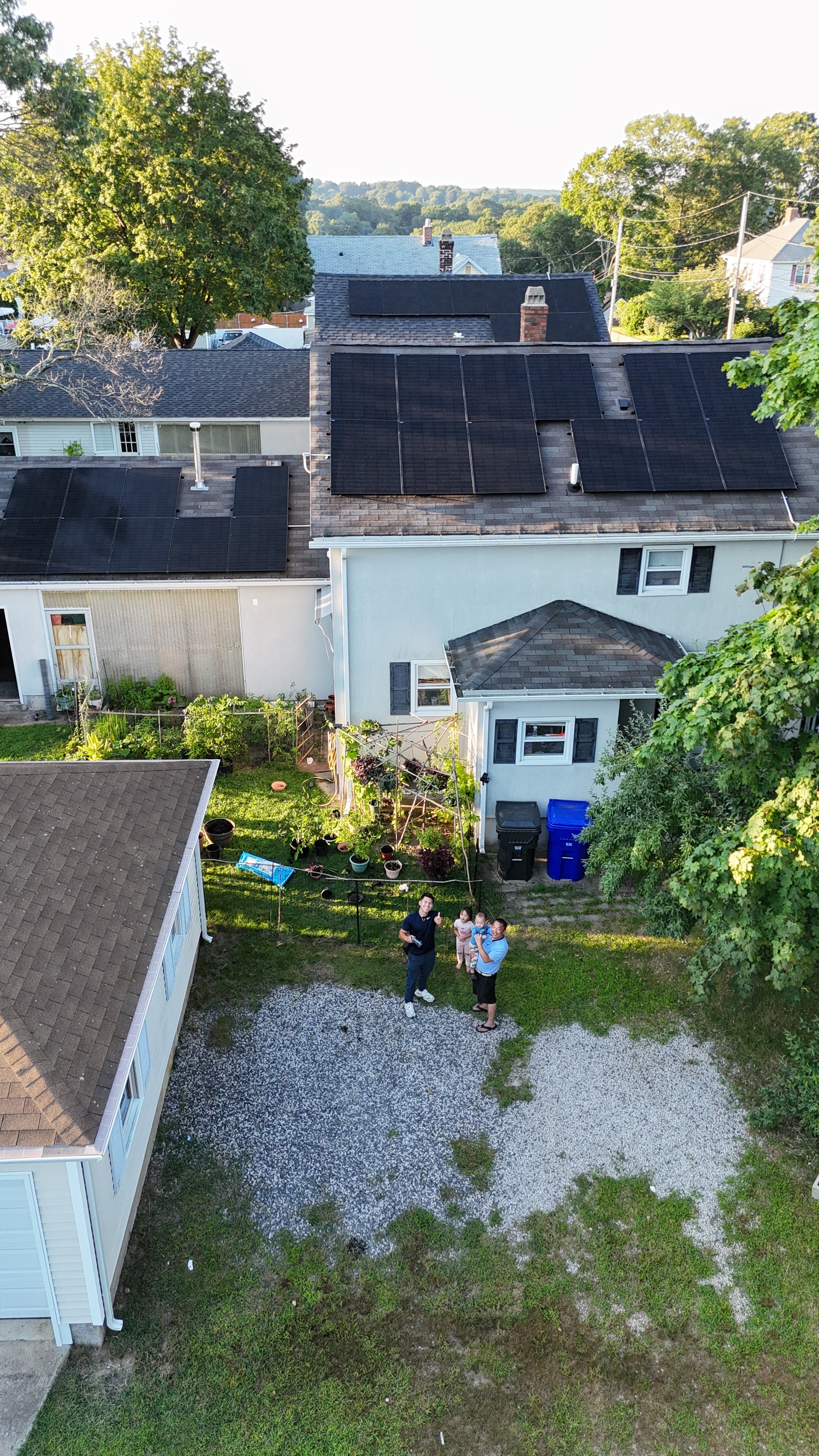 Aerial view of a residential area with a house featuring solar panels on the roof. In the backyard, three adults and a child stand on a gravel area near a garden with several potted plants. Trees and other houses surround the area.