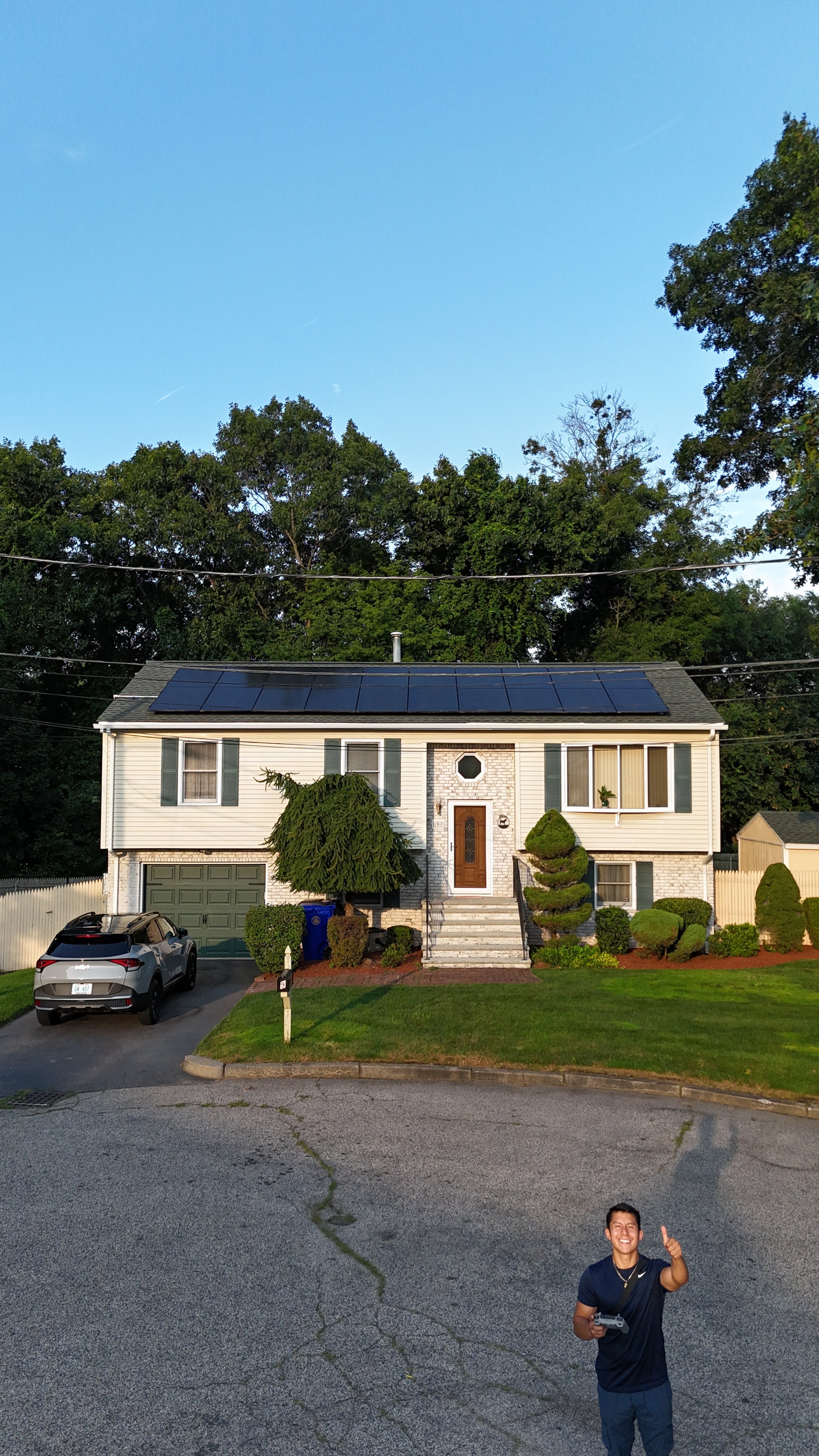 Two-story suburban house with solar panels on the roof, a driveway with an SUV, and a well-kept lawn, surrounded by trees, featuring a person standing in the foreground, smiling and giving a thumbs up.