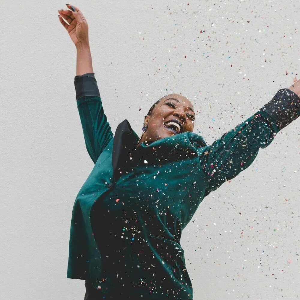 A woman in a teal jacket celebrating her financial clarity with arms raised as colorful confetti falls around her.