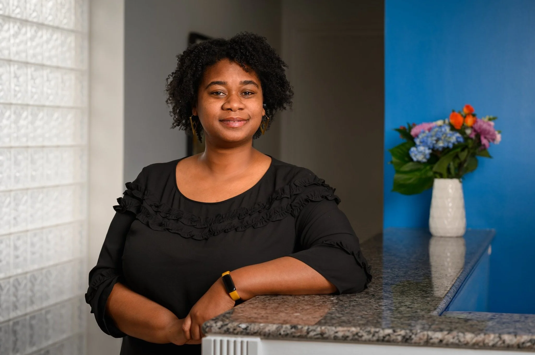 A woman with curly black hair, wearing a black blouse with ruffled details, standing next to a granite counter with her right arm resting on it. There is a white vase with colorful flowers, including pink, purple, blue, and orange blooms, on the counter behind her. The background includes a blue wall and a glass block wall to the left.