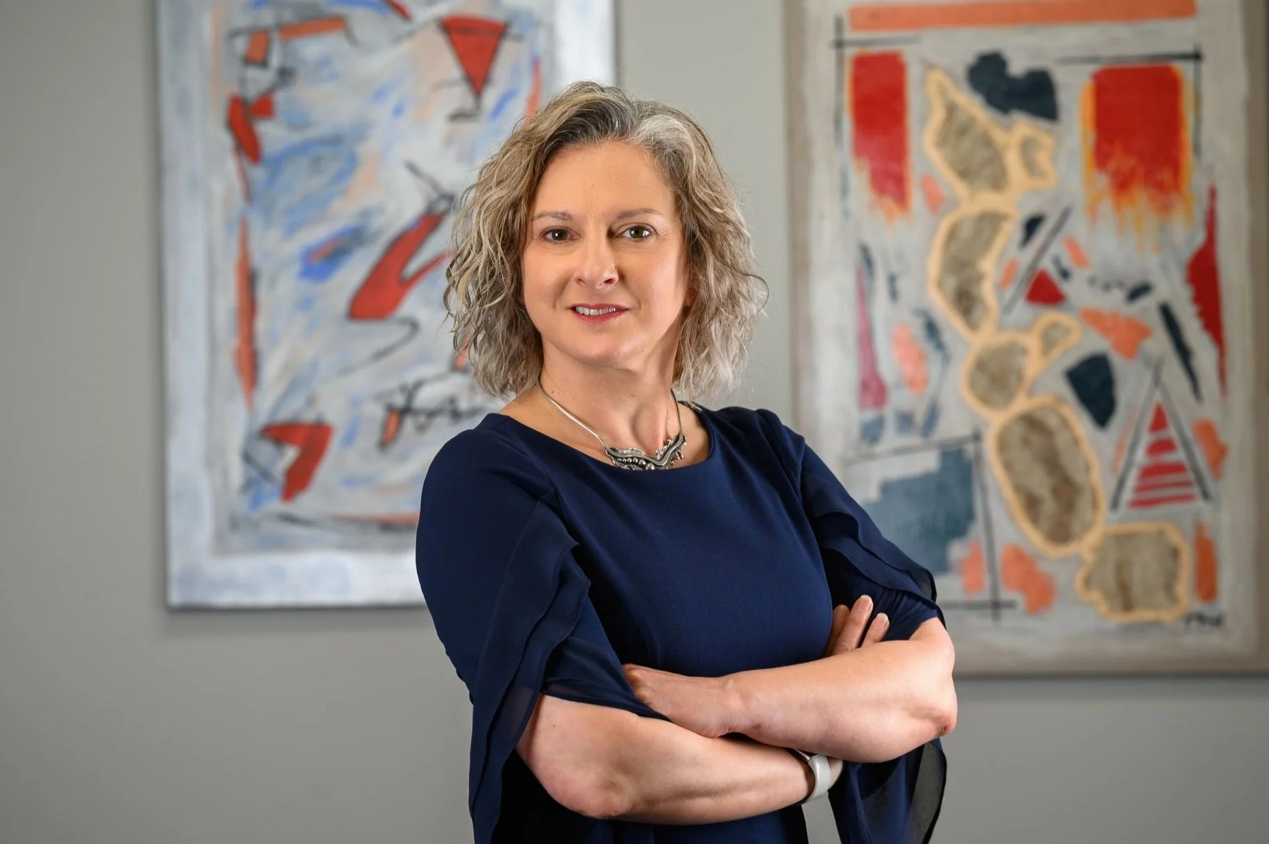 A woman in a dark blue dress standing with her arms crossed in front of abstract artwork on a gray wall.