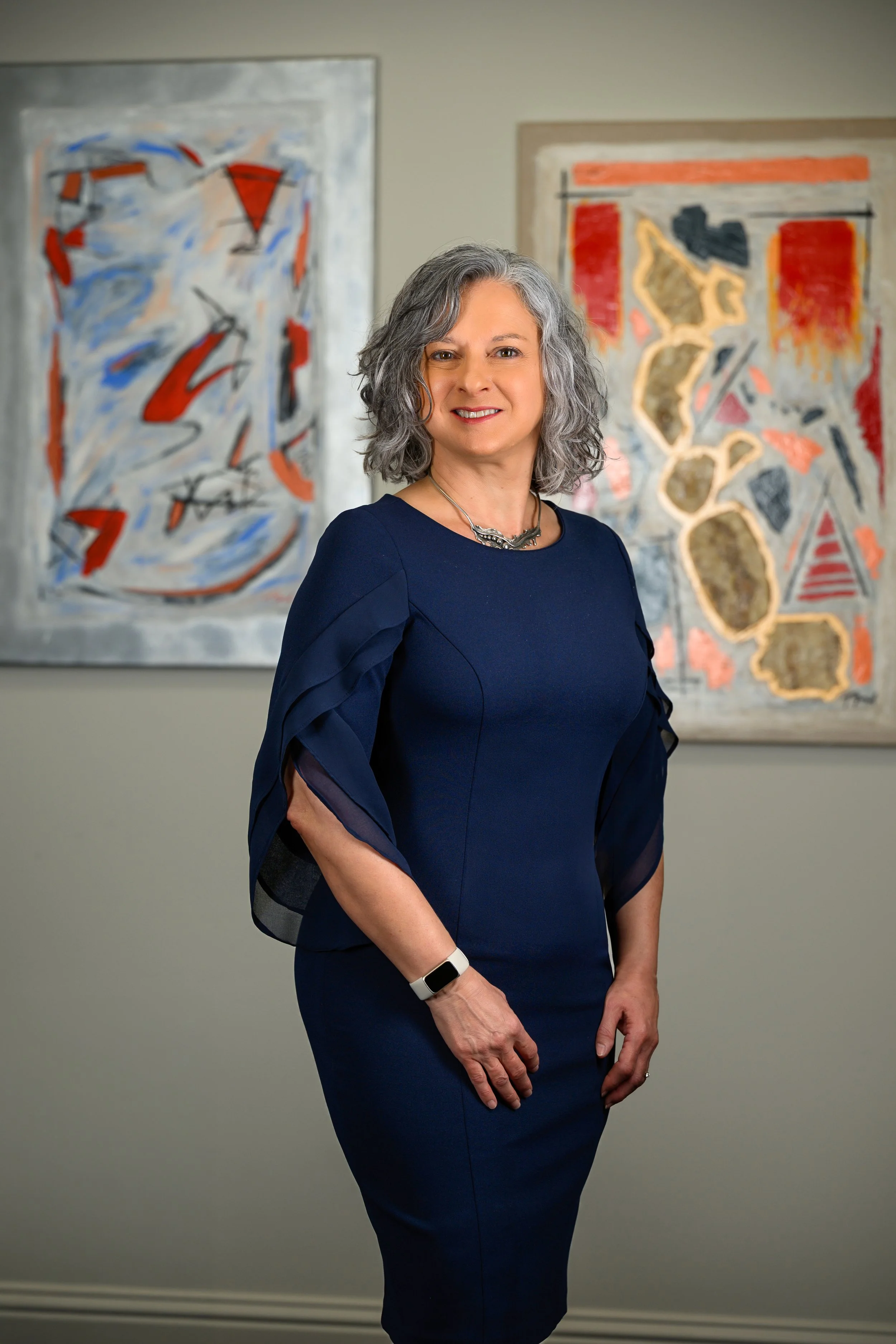 A woman in a dark blue dress standing with her arms crossed in front of abstract artwork on a gray wall.