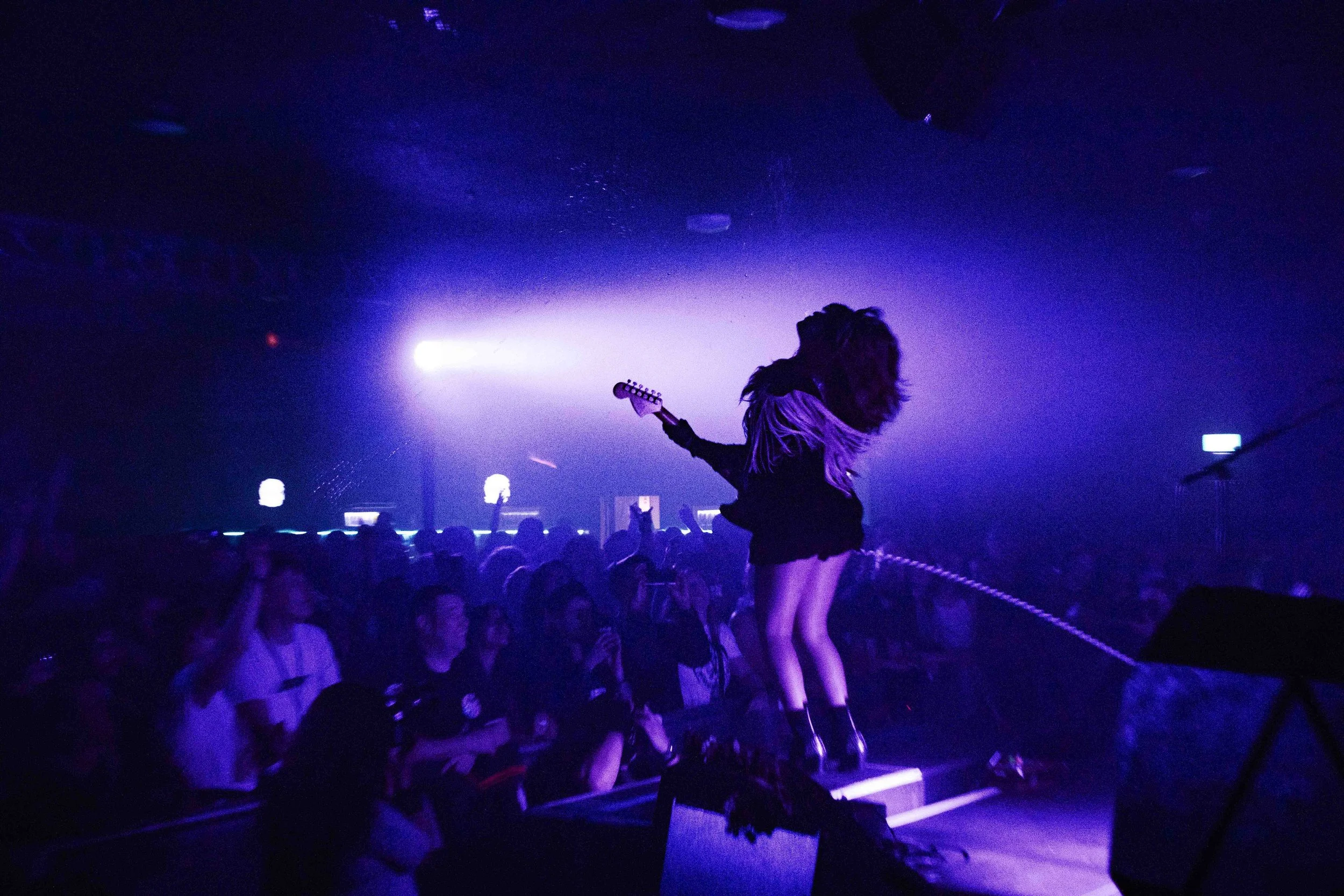A female guitarist performing on stage at a concert, illuminated by purple and blue stage lights, with audience members watching.