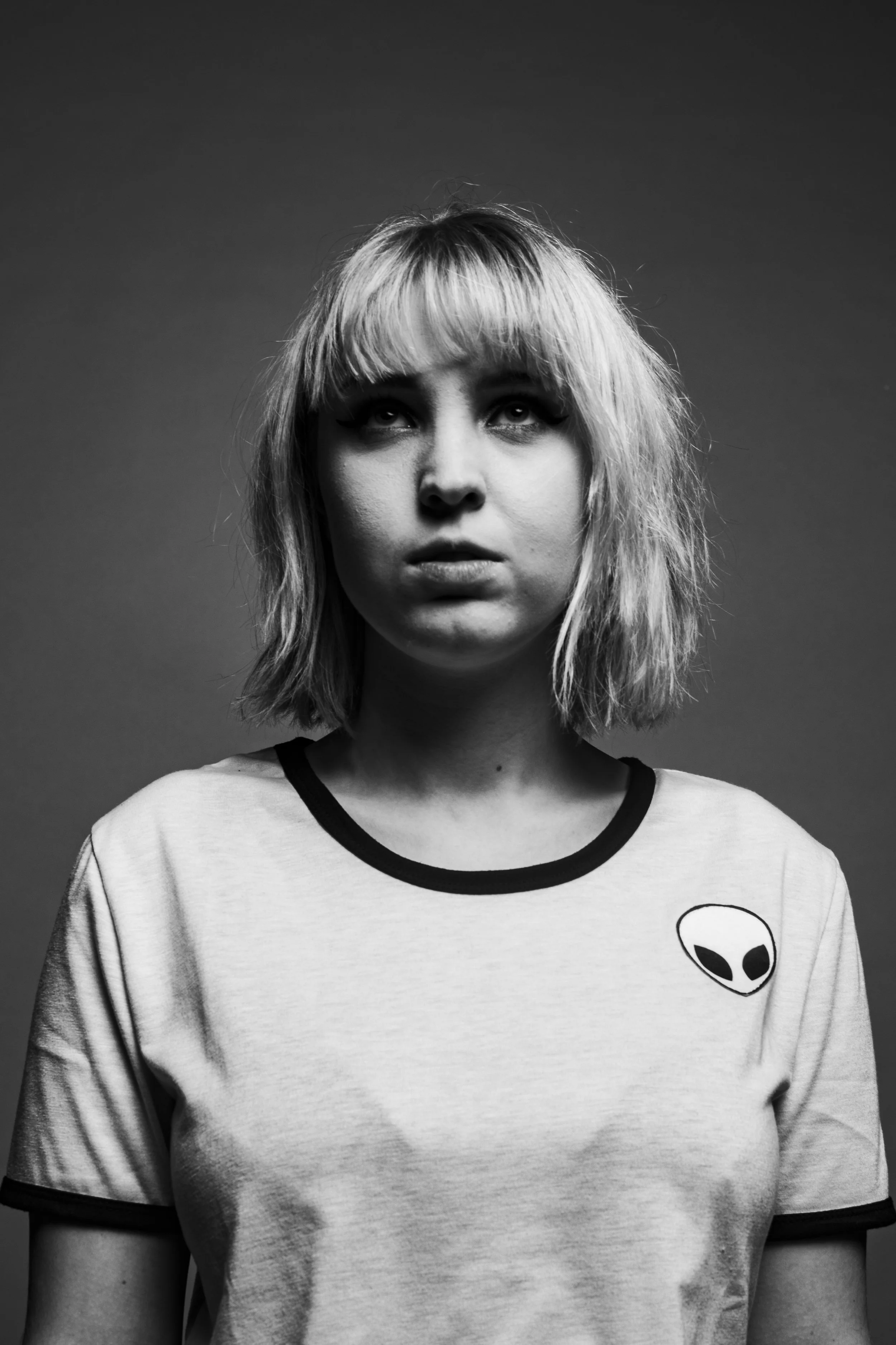 Black and white portrait of a woman with shoulder-length, messy hair and bangs, wearing a t-shirt with an alien head graphic.